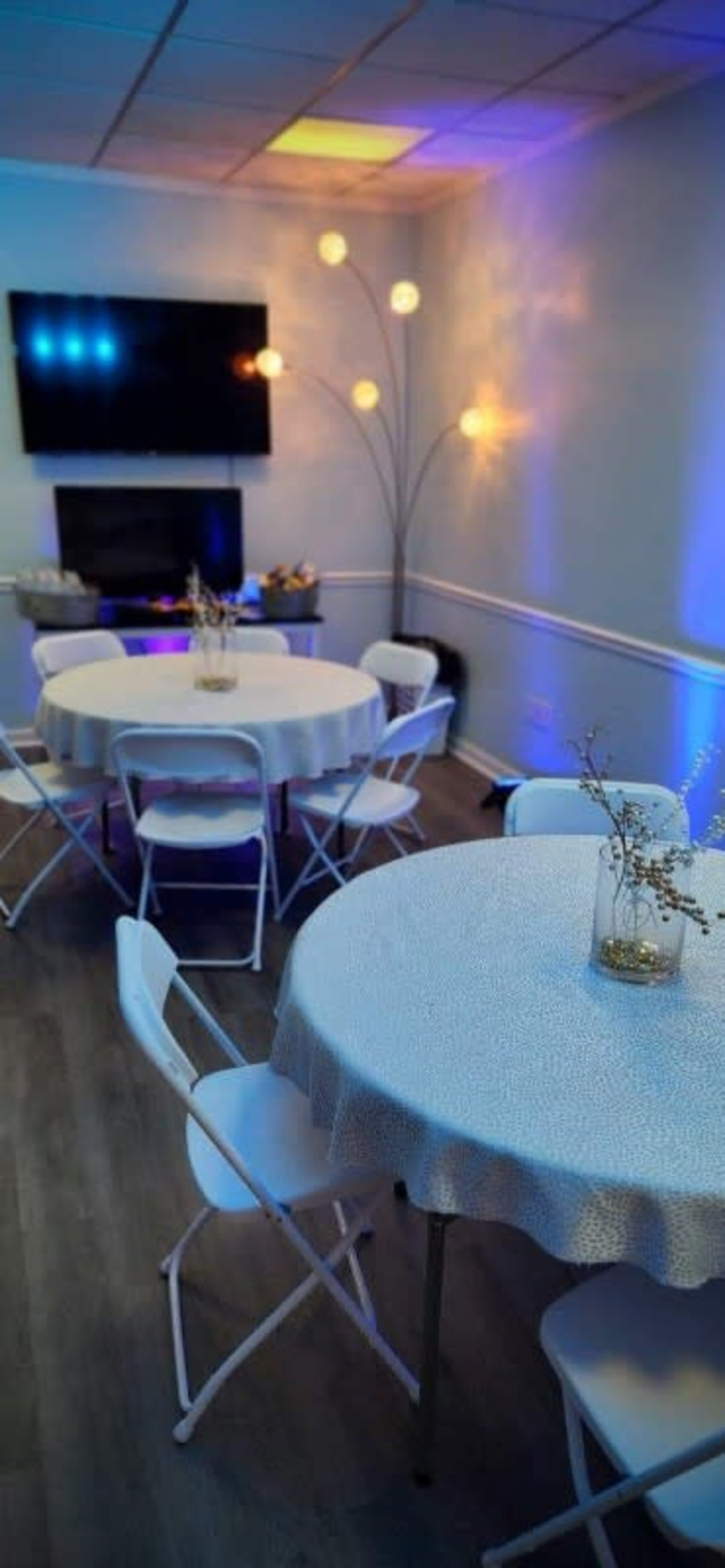 The image shows a small, well-lit room with two round tables covered in white tablecloths, surrounded by white chairs, and a large wall-mounted TV.