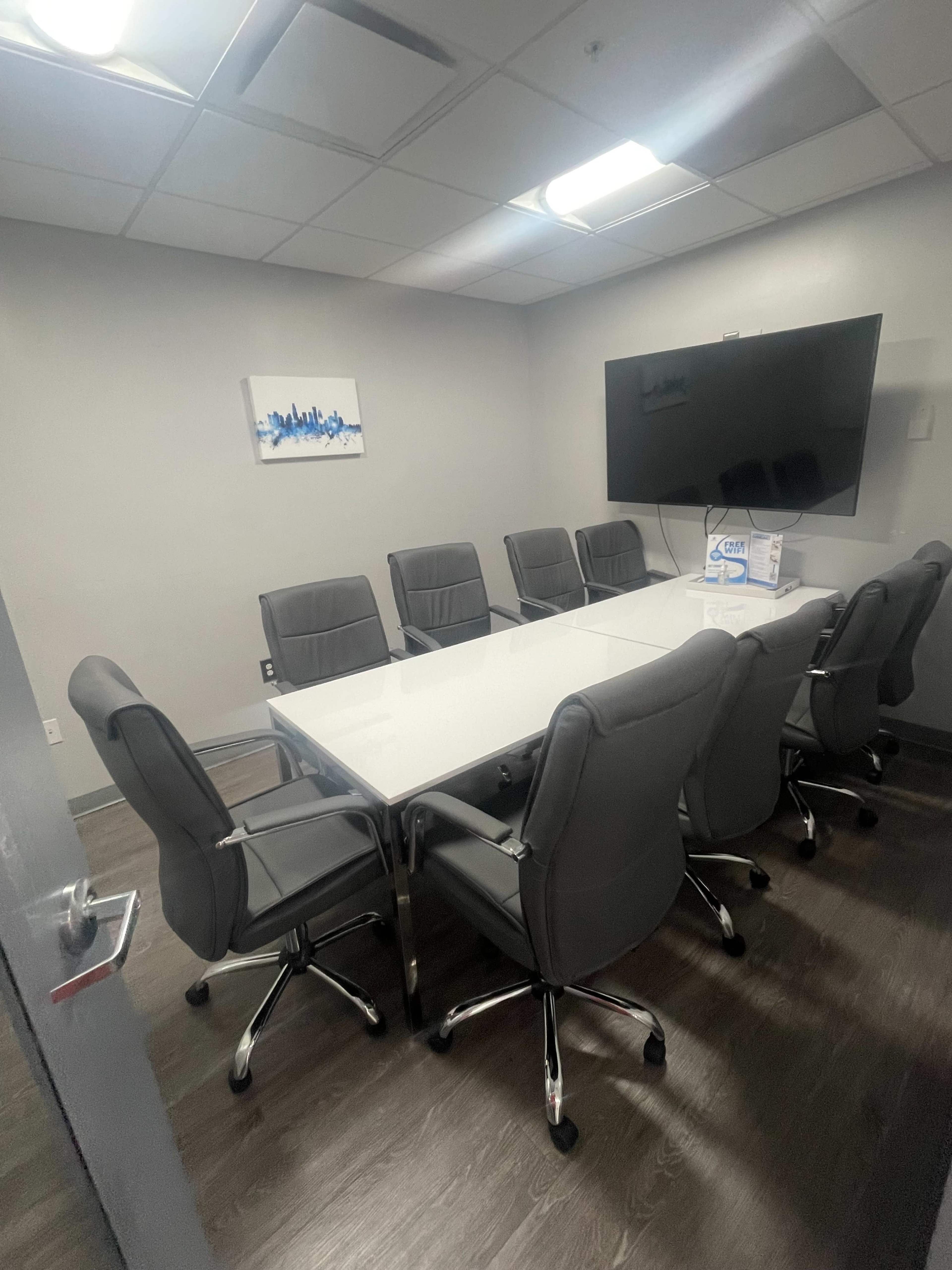The image shows a modern conference room featuring a long glass table surrounded by eight gray ergonomic chairs, with a large screen mounted on the wall and a piece of artwork hanging above.