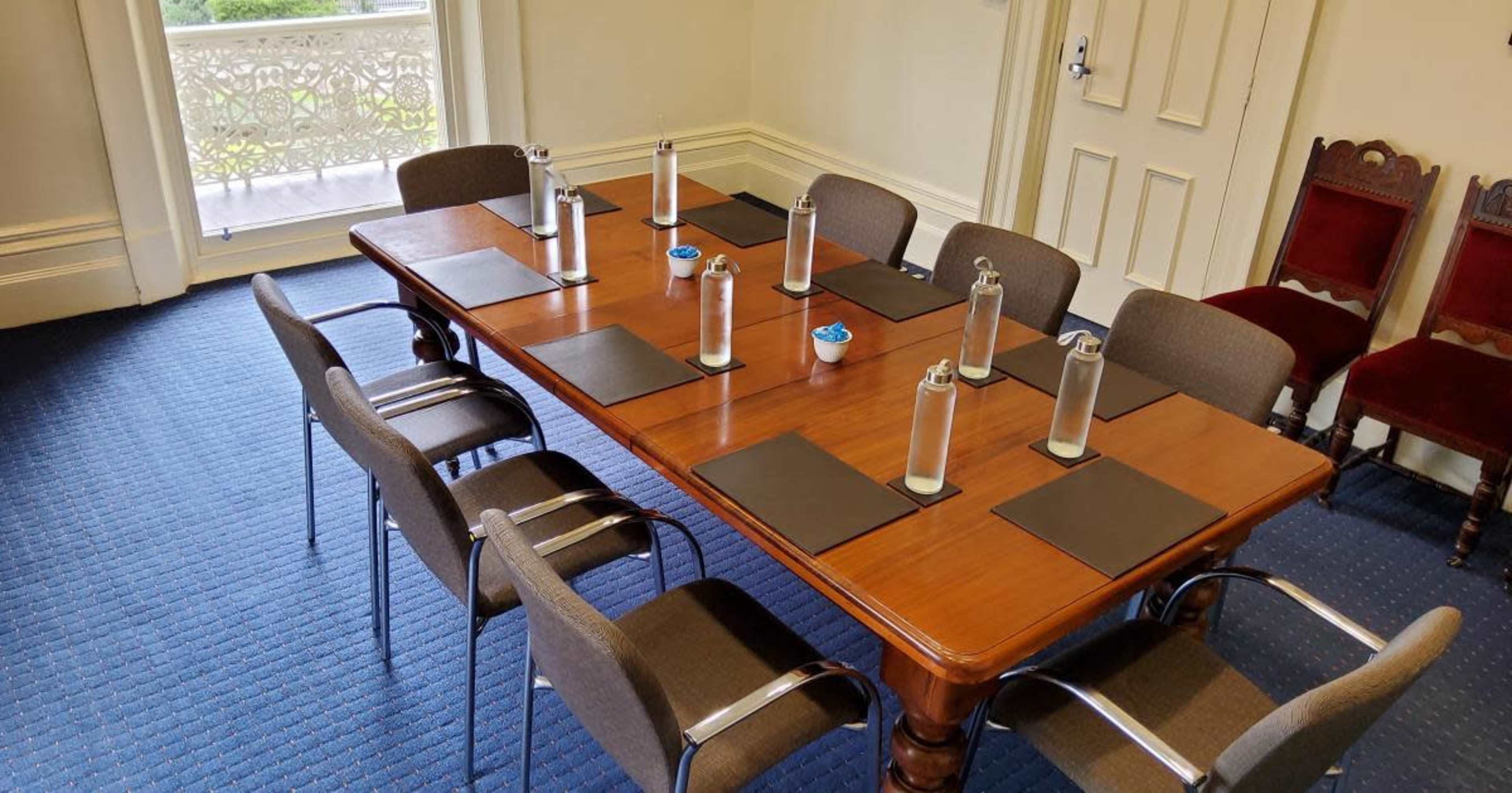 A rectangular wooden conference table is set with placemats, water bottles, and chairs arranged around it in a brightly lit room.