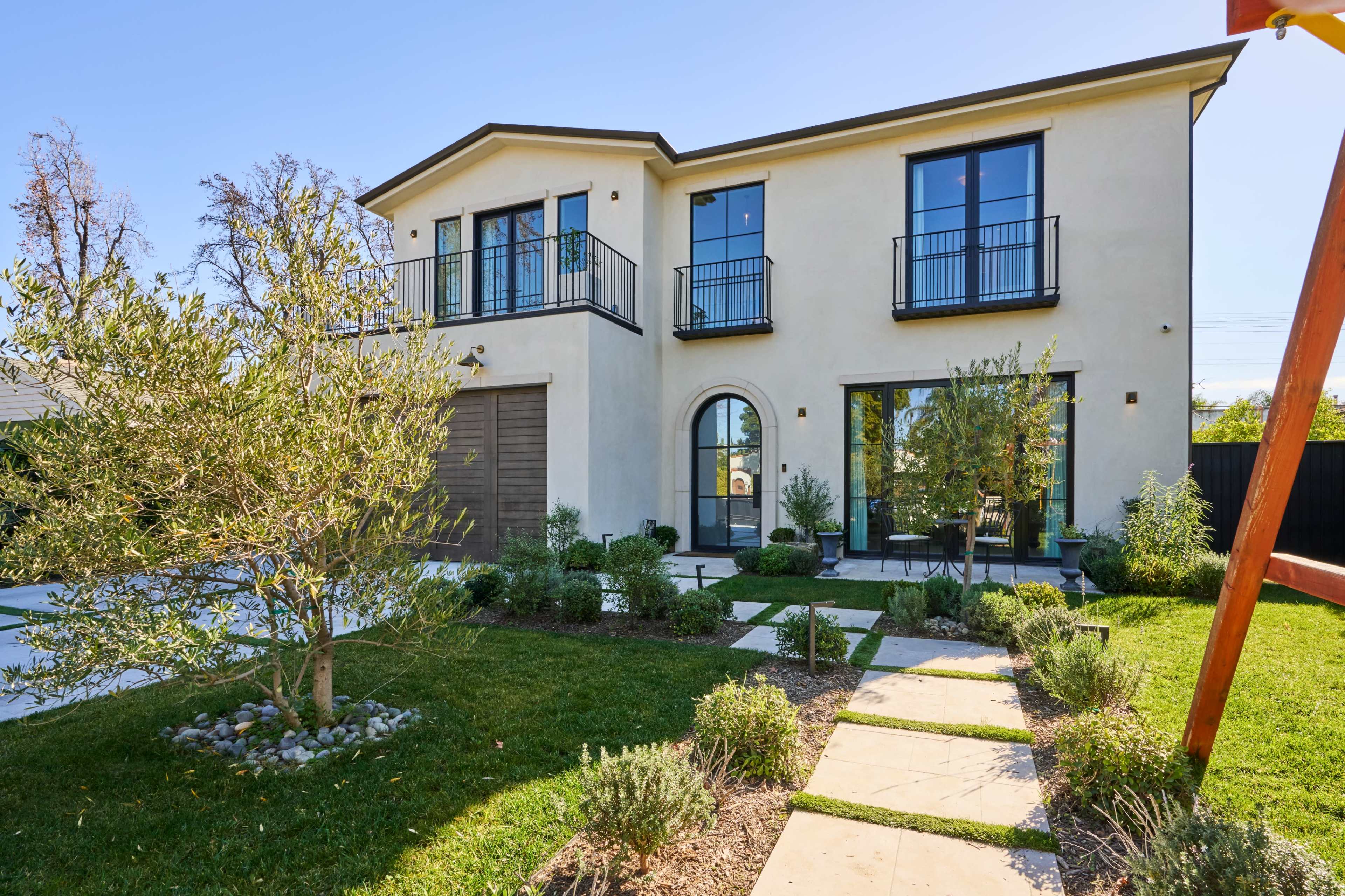 The image shows a modern two-story house with large windows, a brown garage door, and a landscaped front yard featuring pathways and plants.
