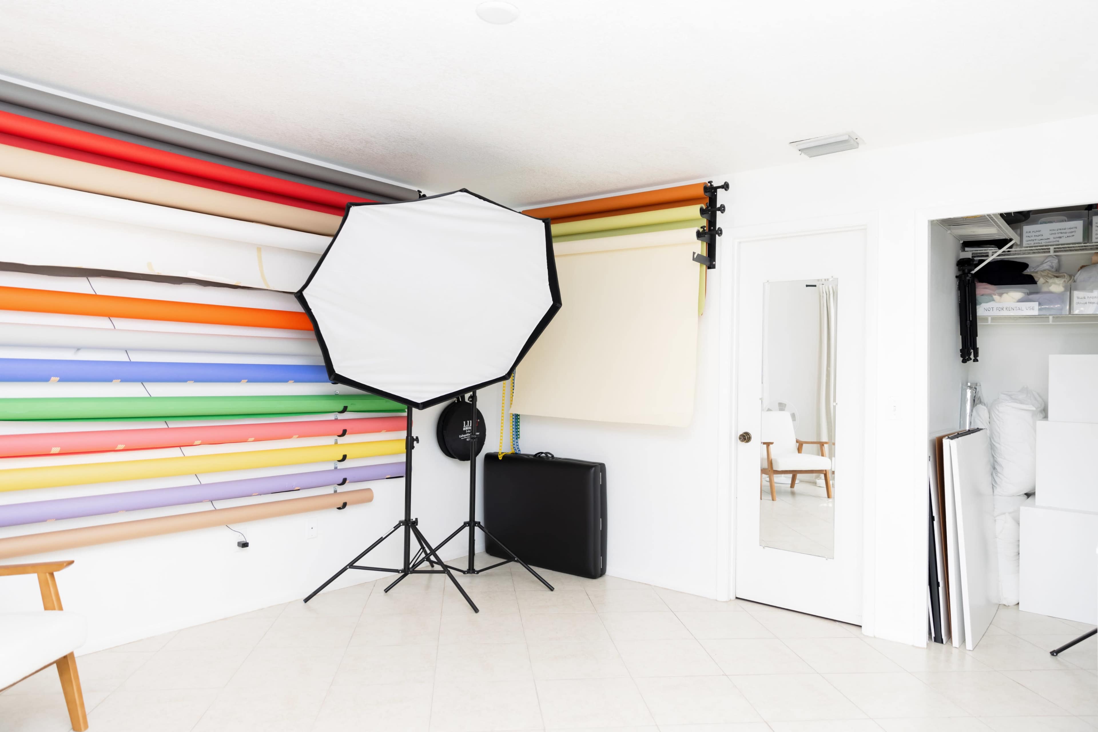 The image shows a photography studio with a wall of colorful backdrop rolls, a large lighting umbrella, and storage space containing white materials.