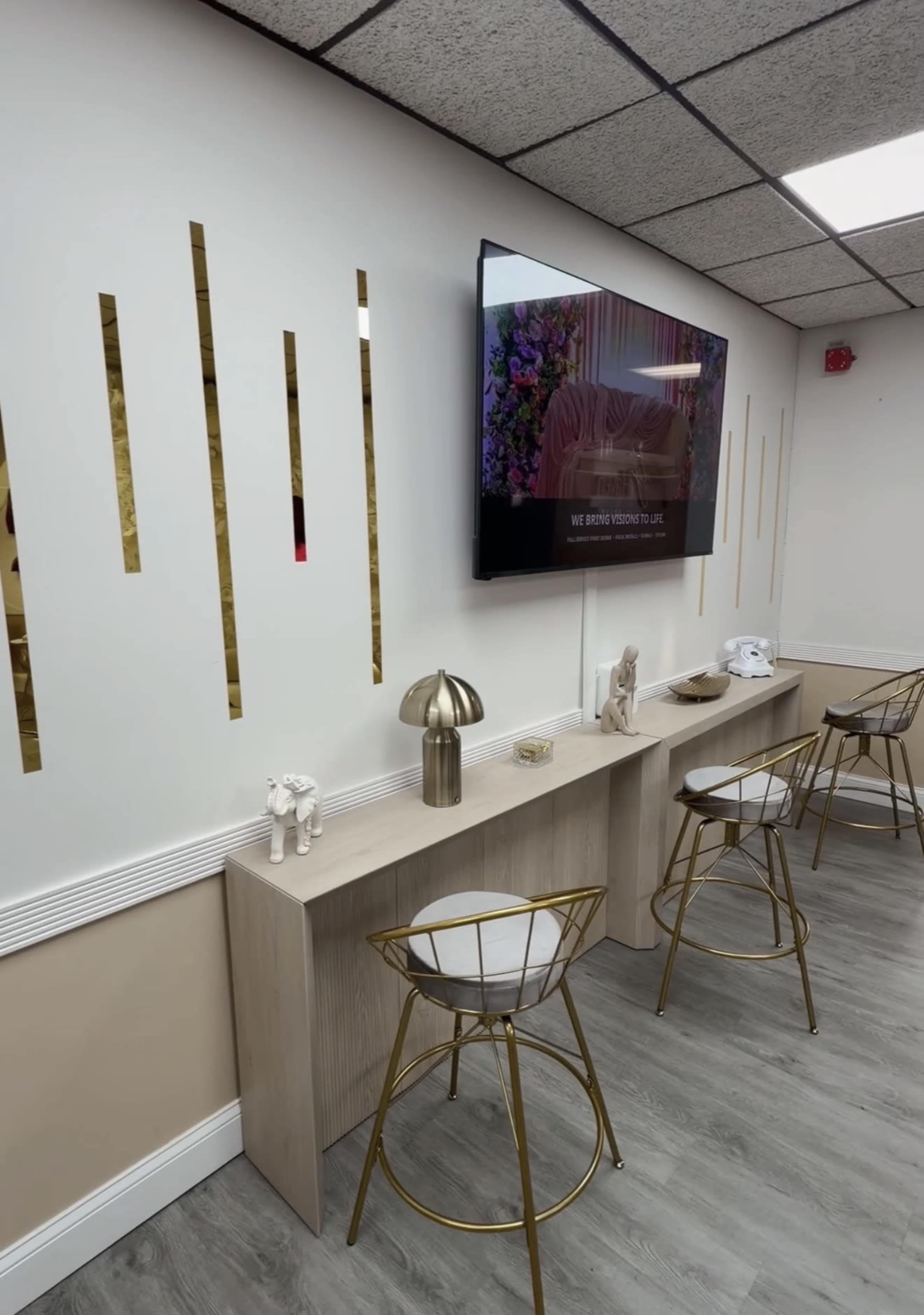 The image shows a modern interior with a light-colored wall featuring vertical gold accents, a flat-screen TV displaying colorful visuals, and a minimalist table with decorative items and gold-accented chairs.