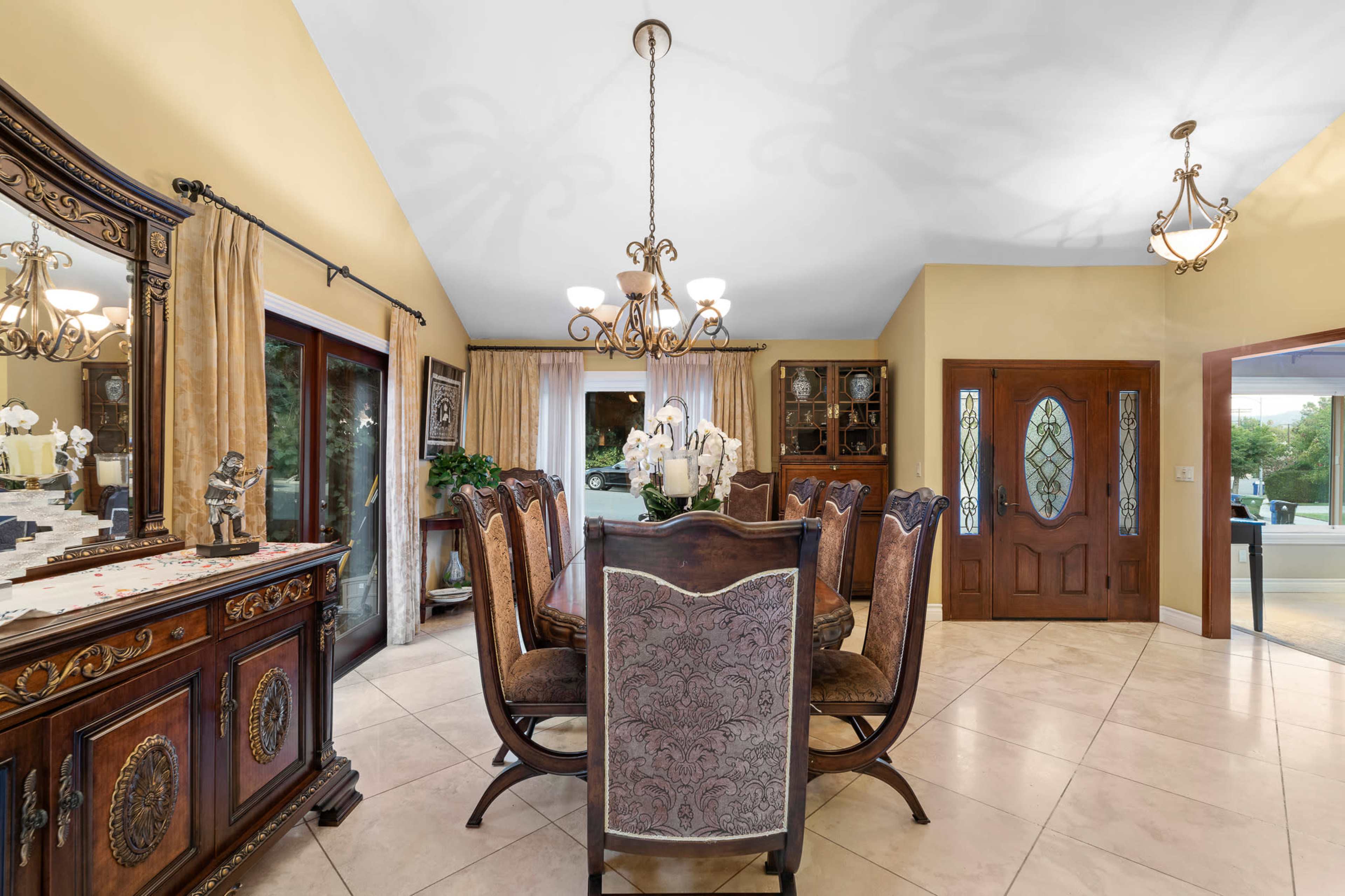 The image shows a dining room with a wooden dining table surrounded by upholstered chairs, a decorative sideboard, and large windows allowing natural light.