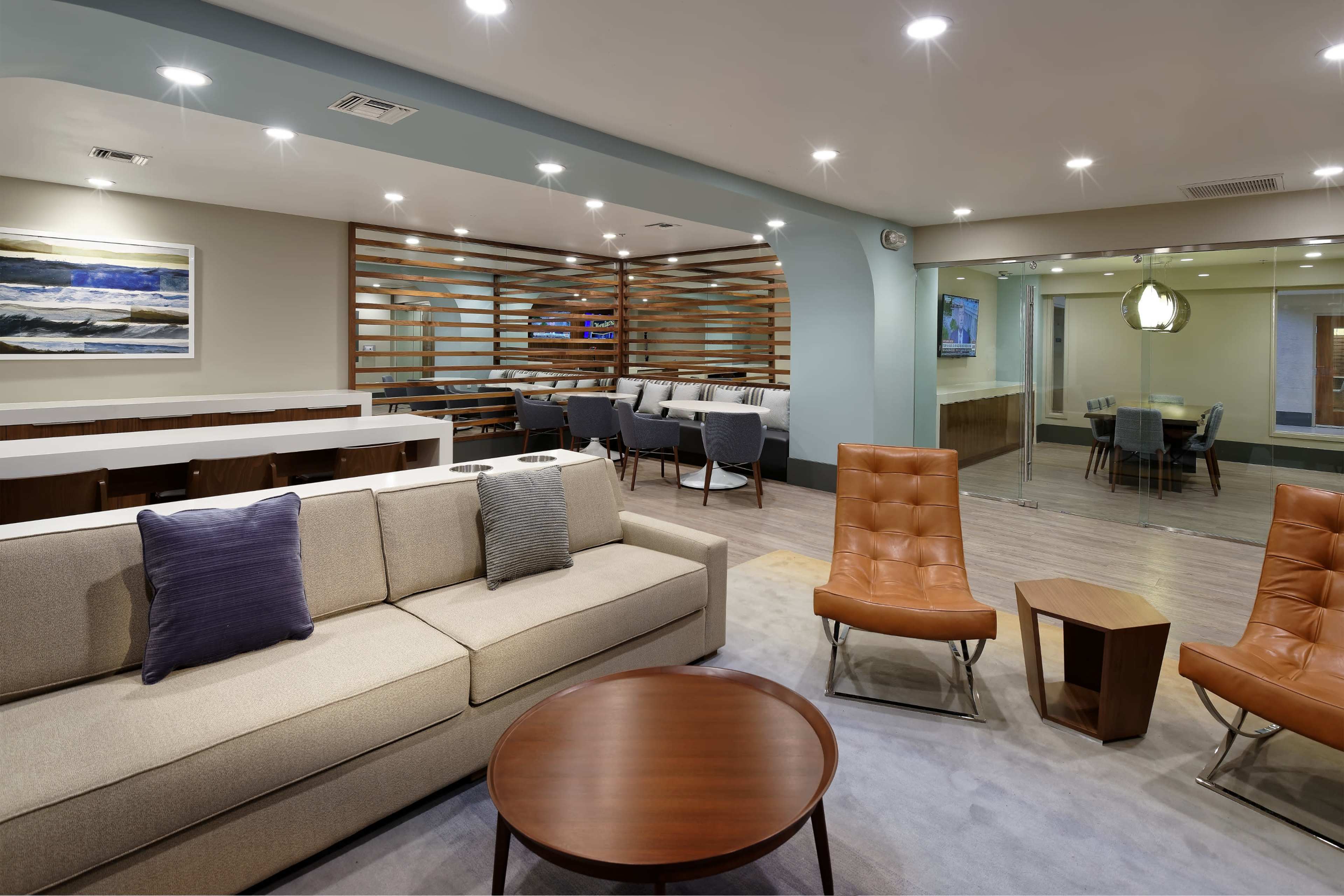 A modern lounge area with a light-colored sofa, two orange chairs, a round coffee table, and a dining area in the background featuring wooden accents and glass walls.