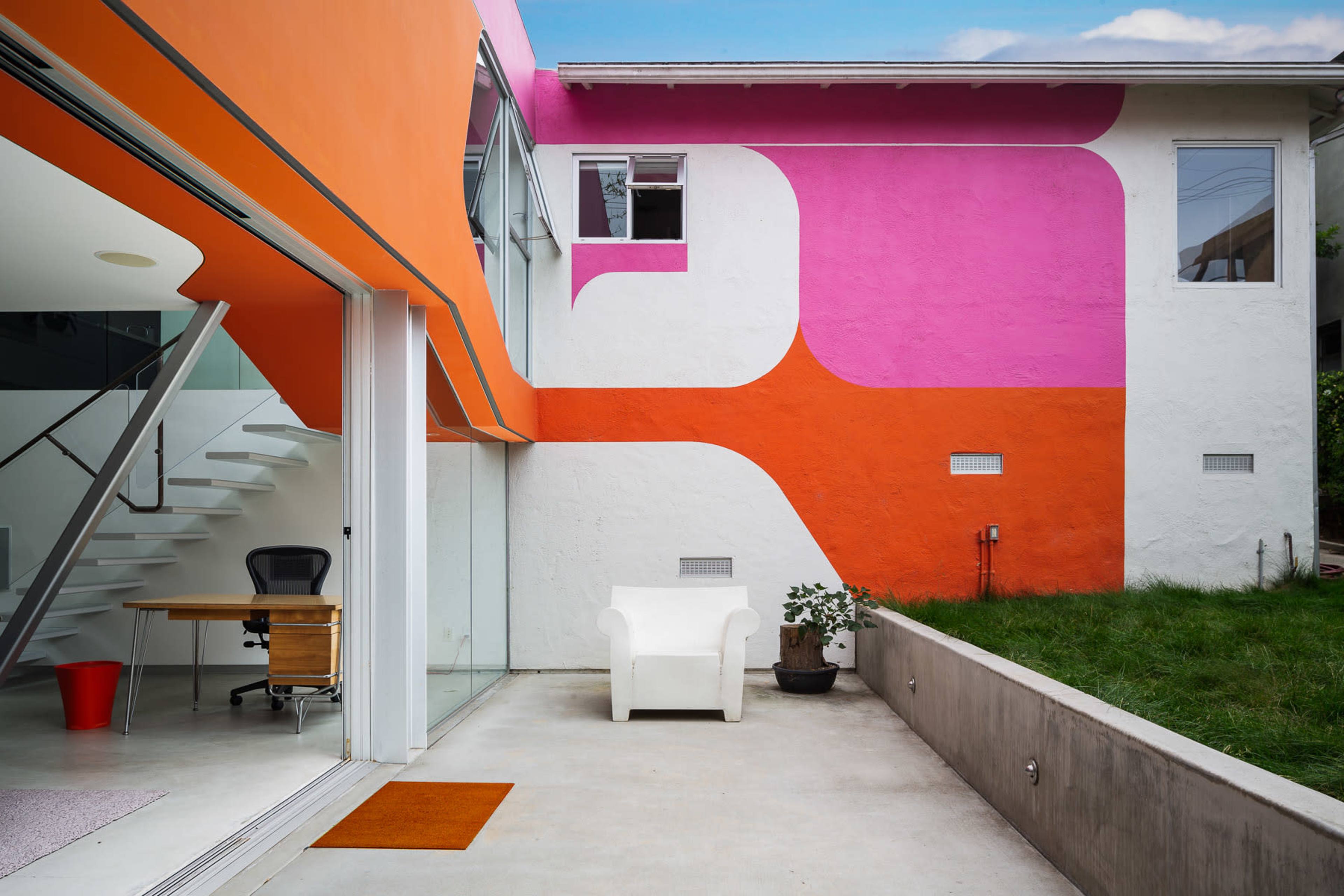 A modern house features a colorful exterior with orange and pink shapes on the walls, and a minimalist white chair sits in the concrete yard.