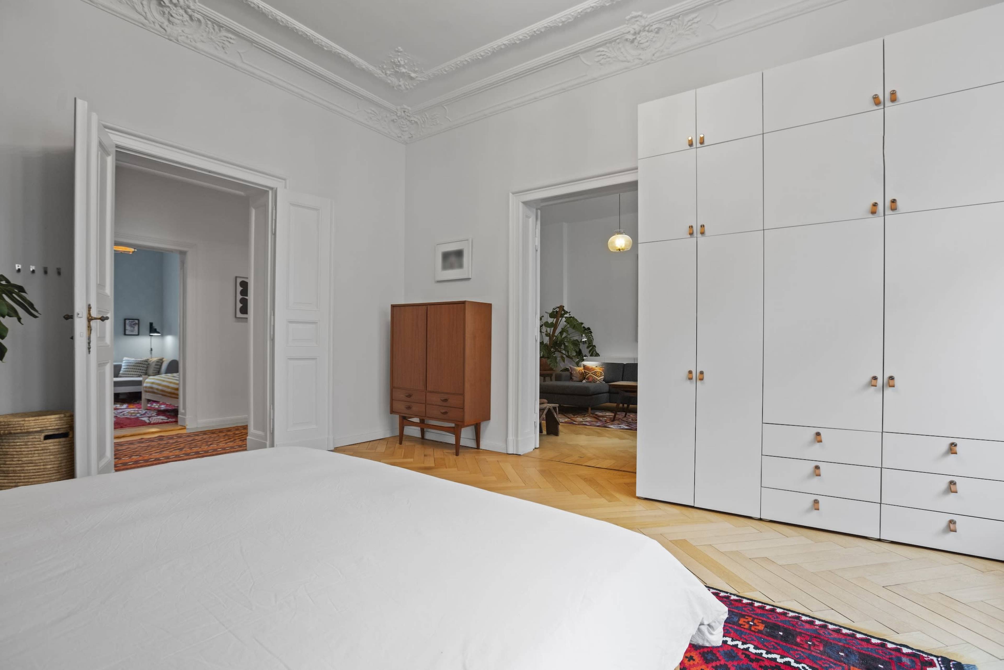 The image shows a bedroom featuring a white bedspread, a wooden wardrobe, and a light-colored dresser, with a patterned rug on the herringbone wooden floor.