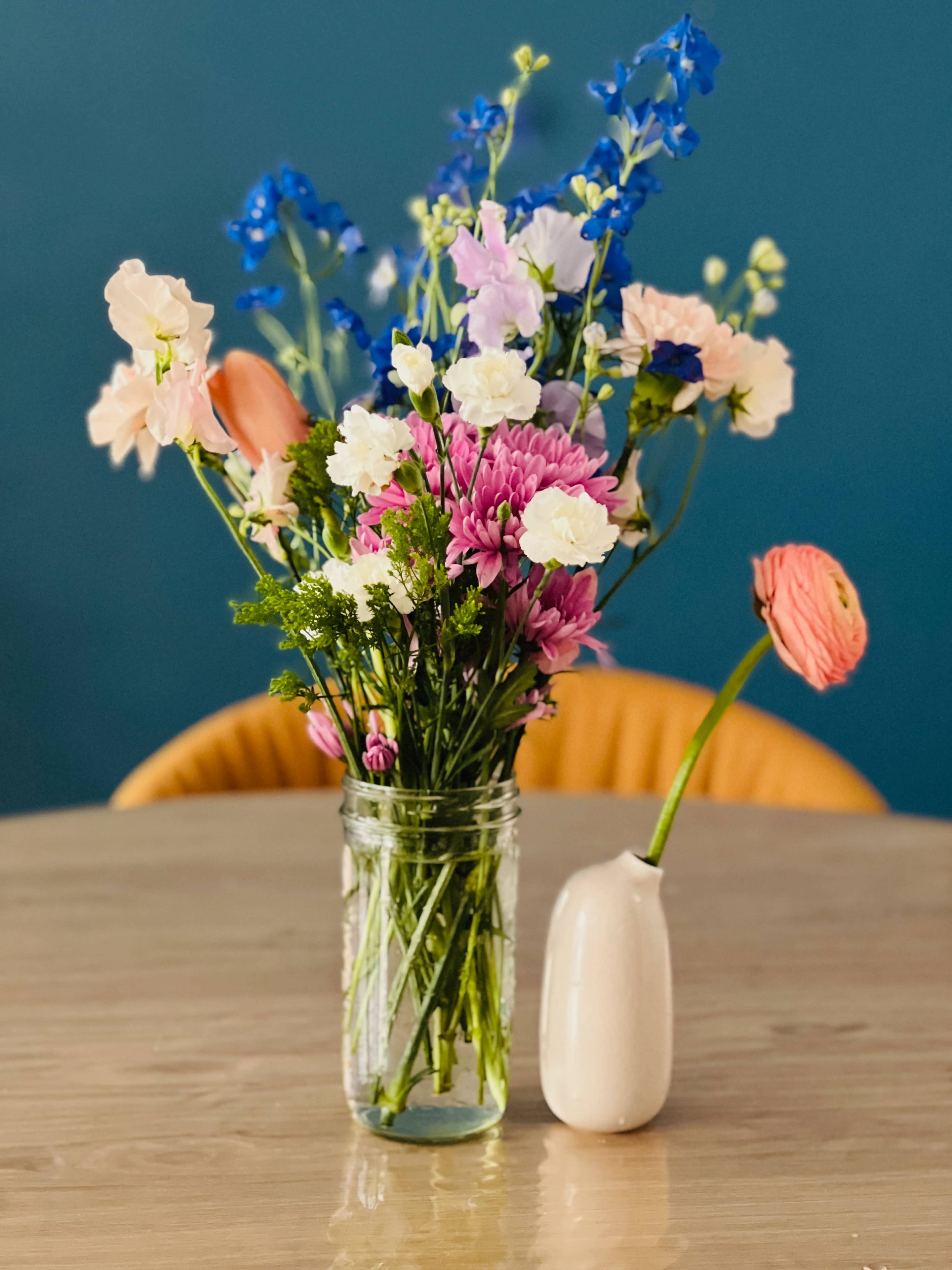 A colorful bouquet of flowers is arranged in a clear jar and a small ceramic vase on a wooden table.
