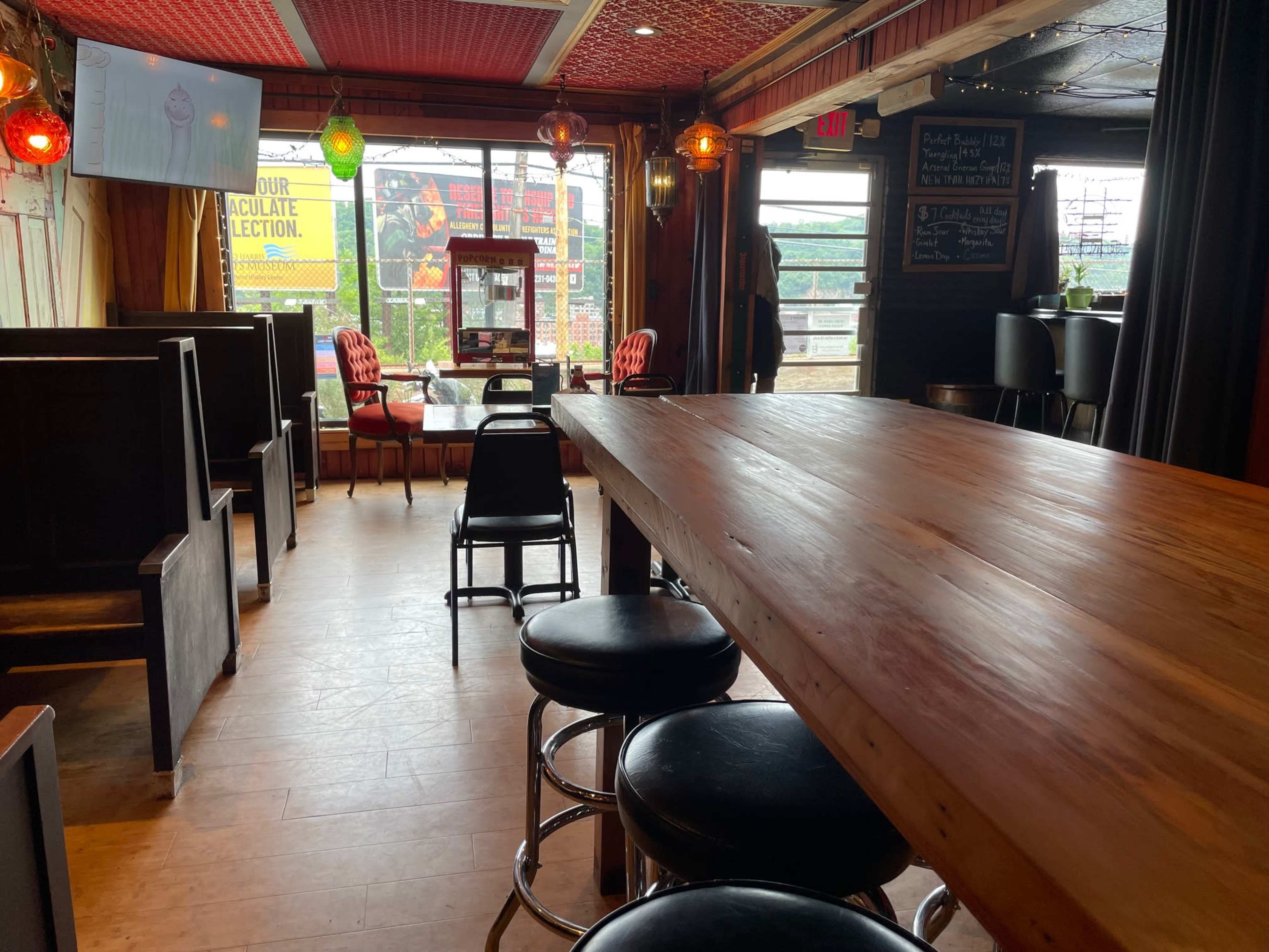 The image shows the interior of a restaurant or bar with wooden tables, black stools, and large windows displaying colorful signage.