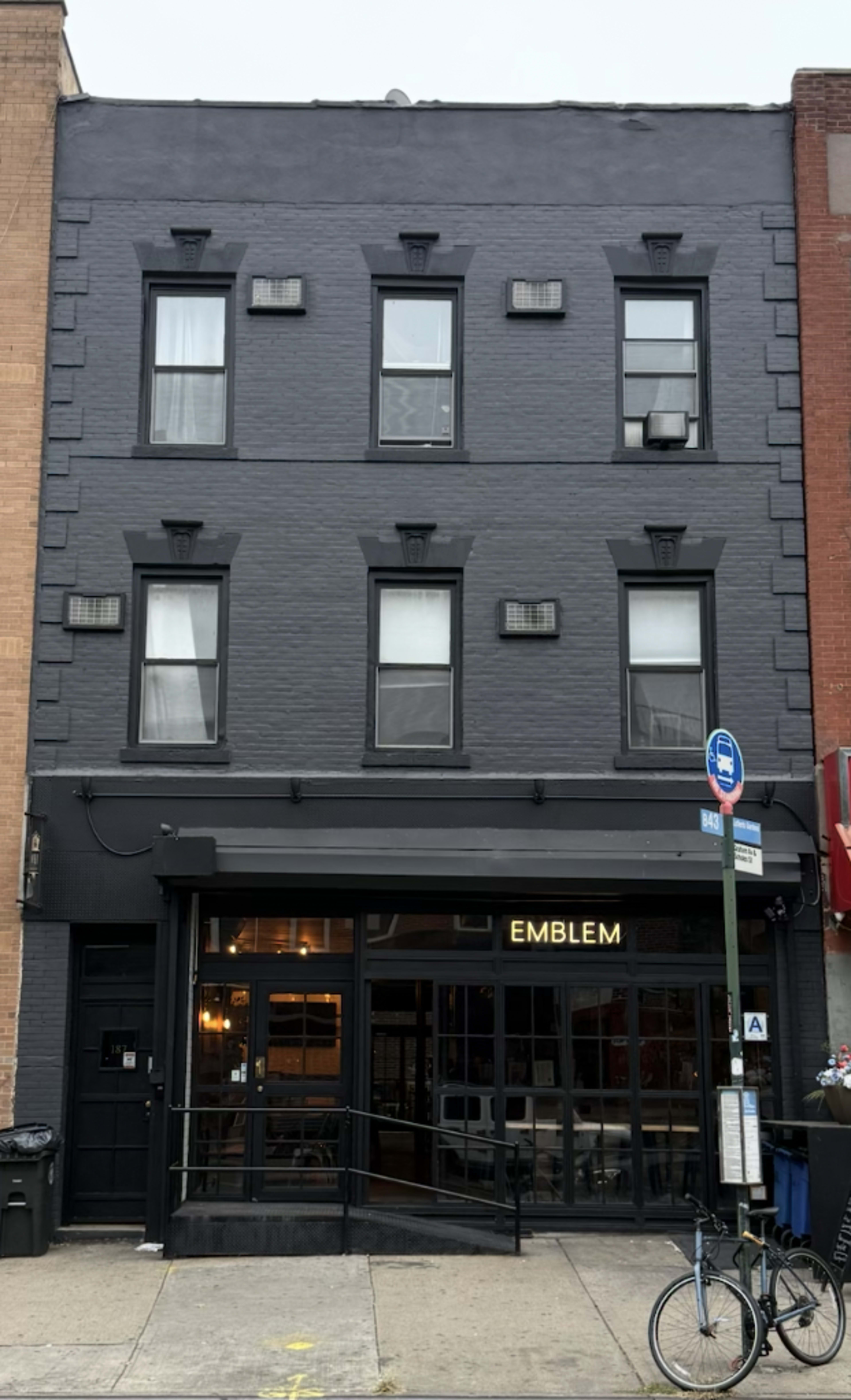 The image shows a three-story gray building featuring large windows and a storefront with the name "EMBLEM" illuminated above the entrance.