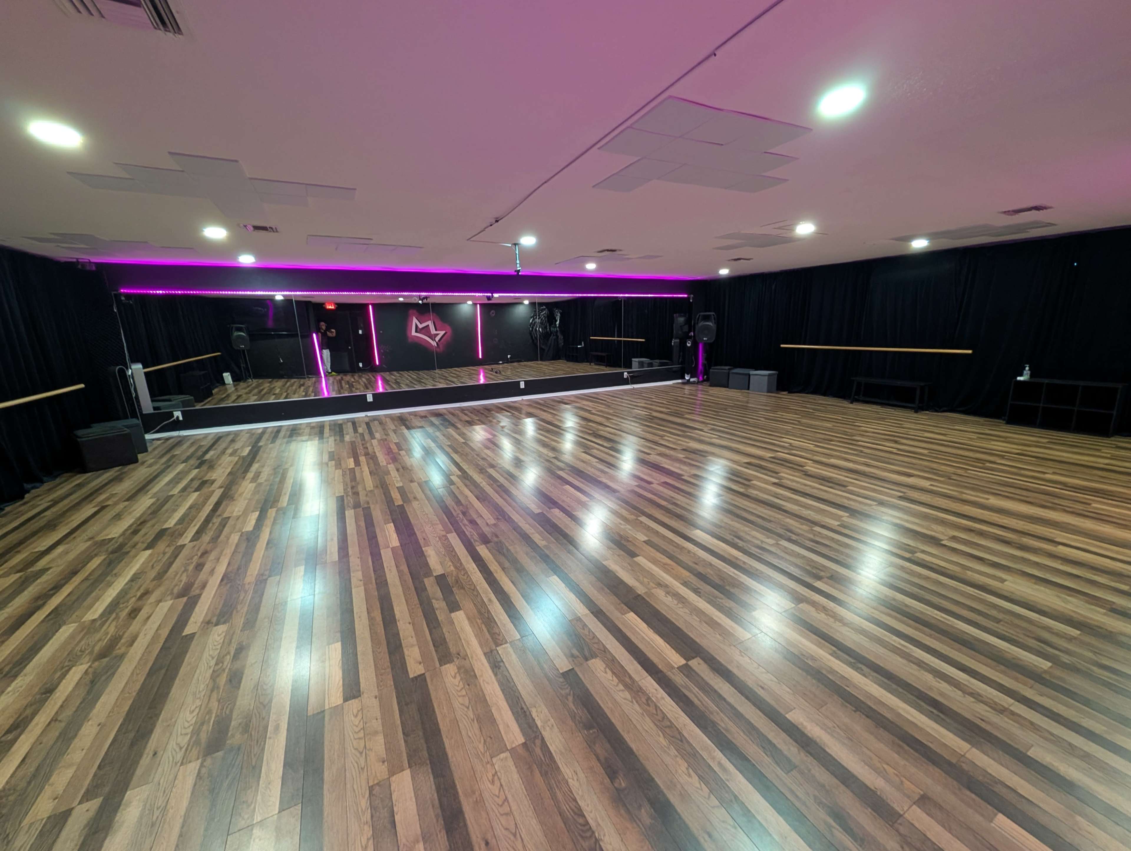 The image shows a spacious dance studio with a polished wooden floor, mirrors along one wall, and soft pink lighting.