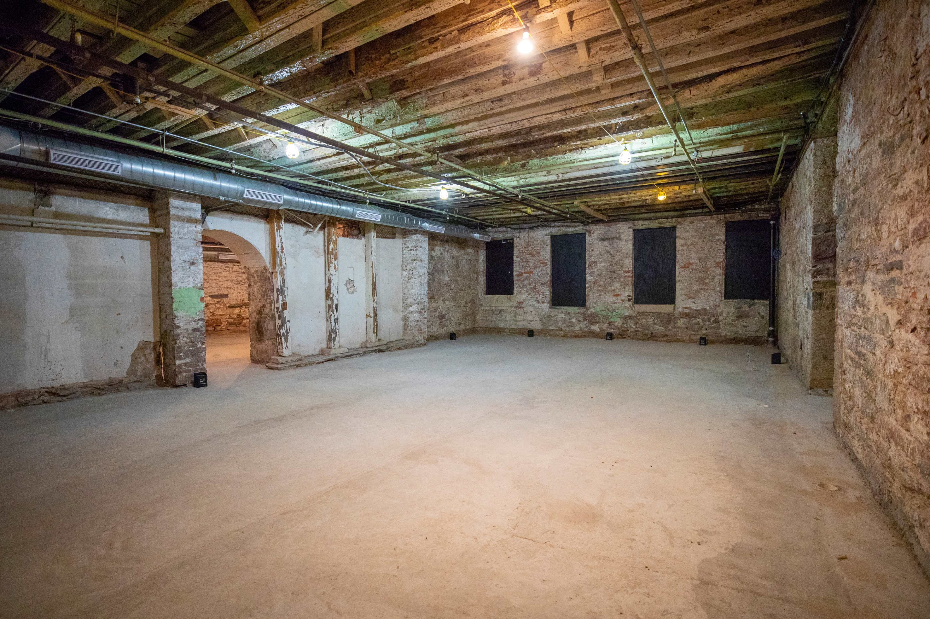 The image shows a spacious, empty basement with exposed wooden beams and stone walls, illuminated by overhead lights.