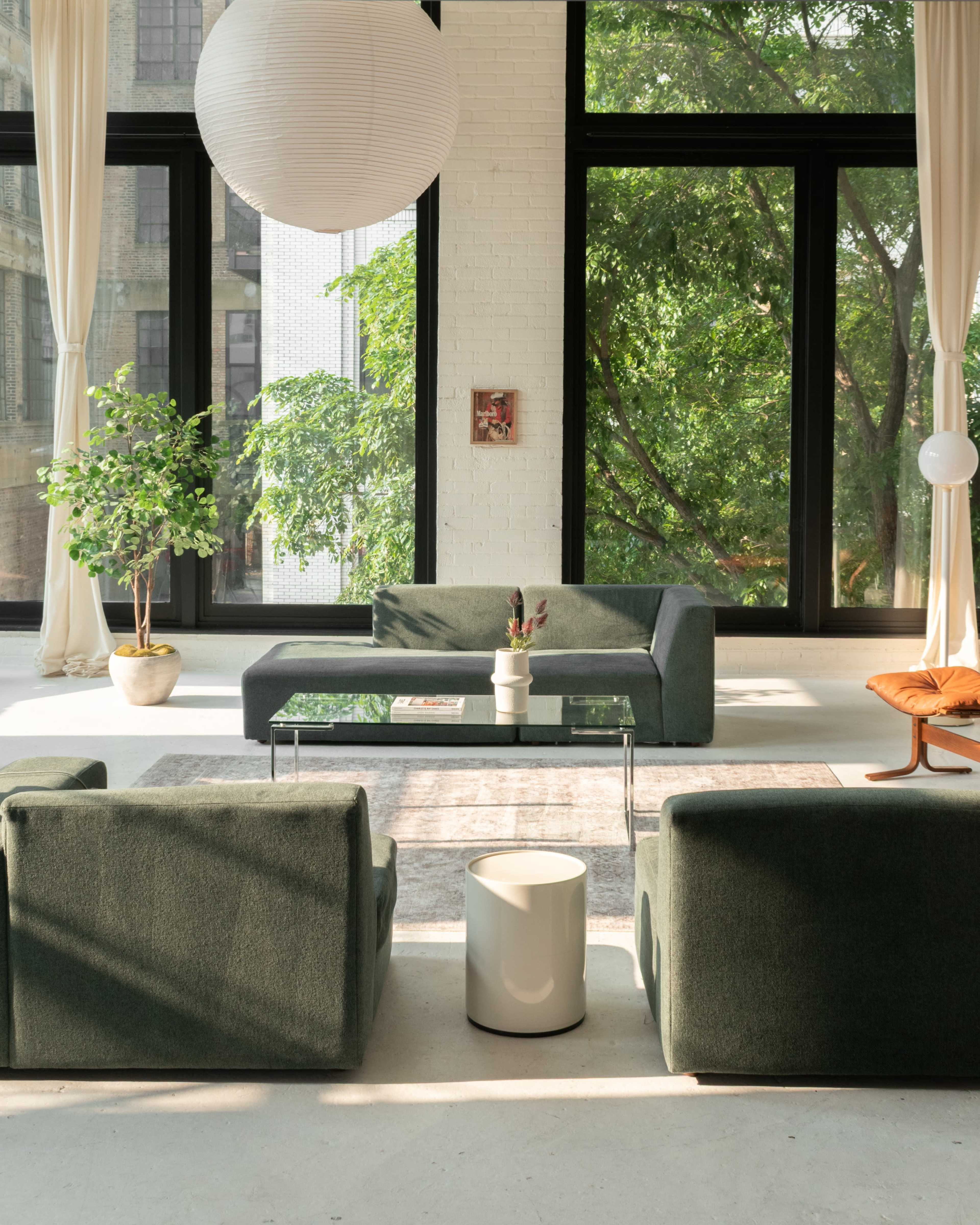 The image shows a bright, modern living room with large windows, green sofas, a glass coffee table, and a potted plant.