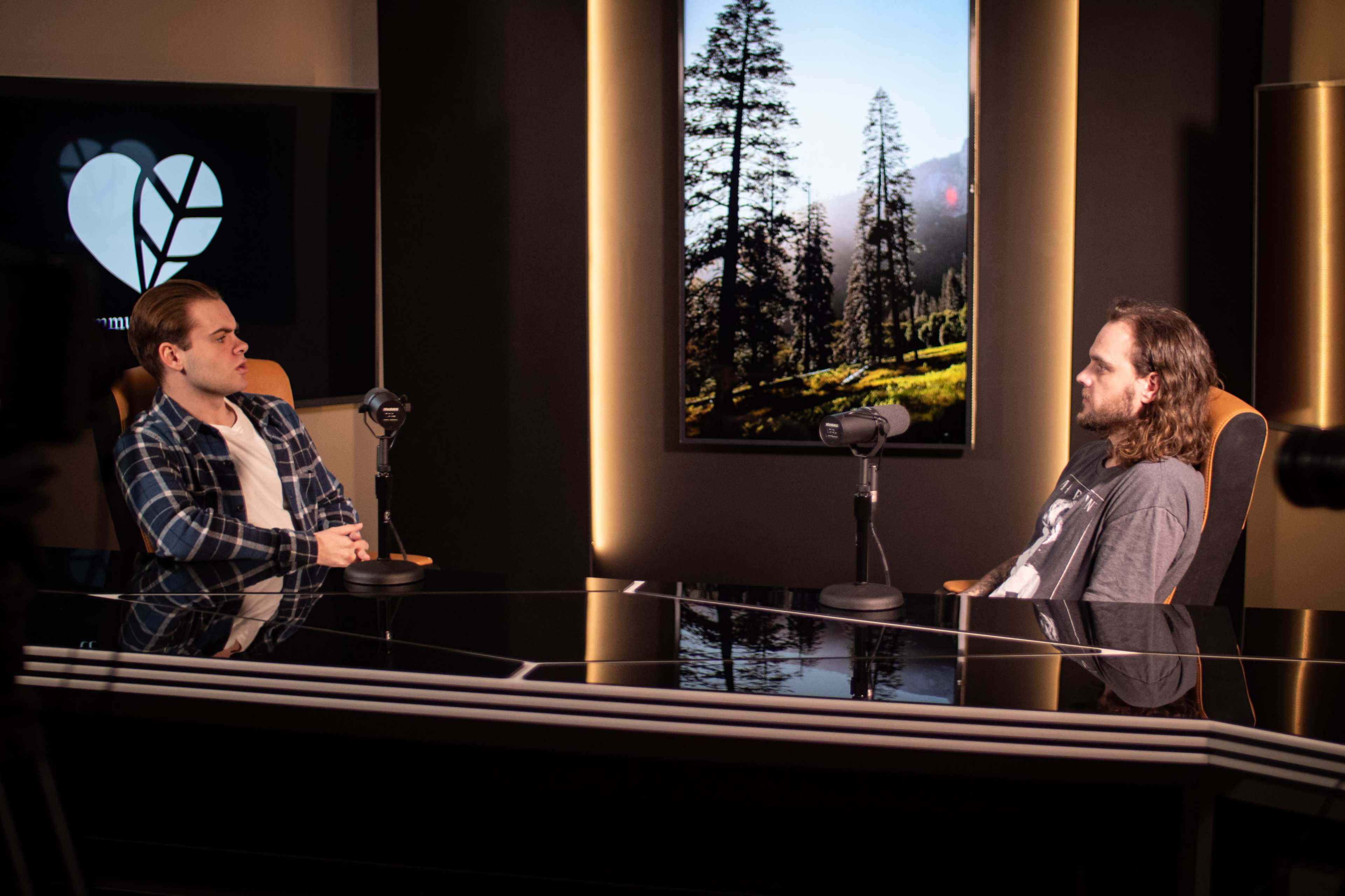 Two men sit across from each other at a glossy table, each in front of a microphone, with a large nature-themed backdrop visible behind them.
