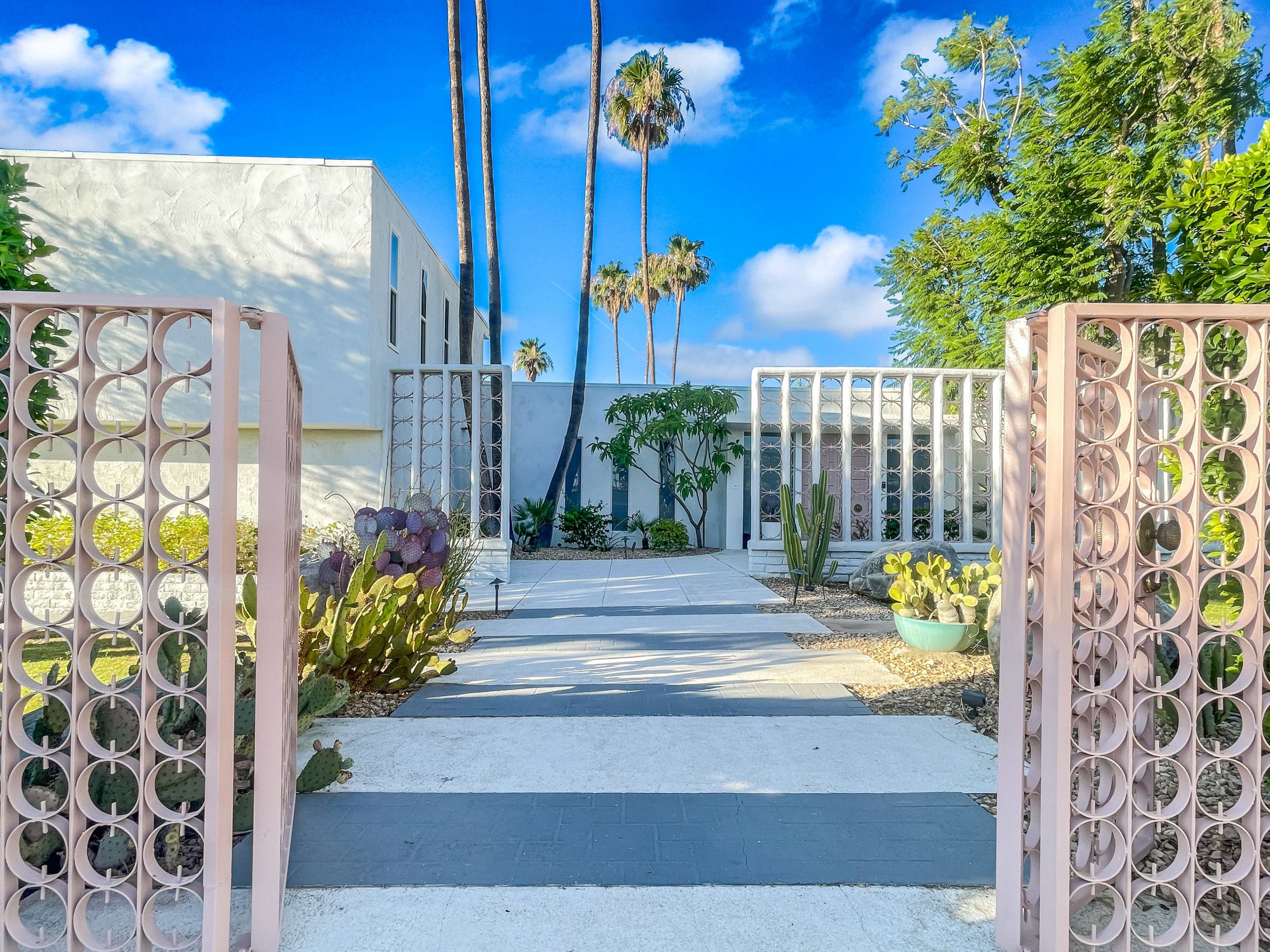 A pathway lined with greenery and modern architecture leads to a serene entrance flanked by decorative gates and palm trees.