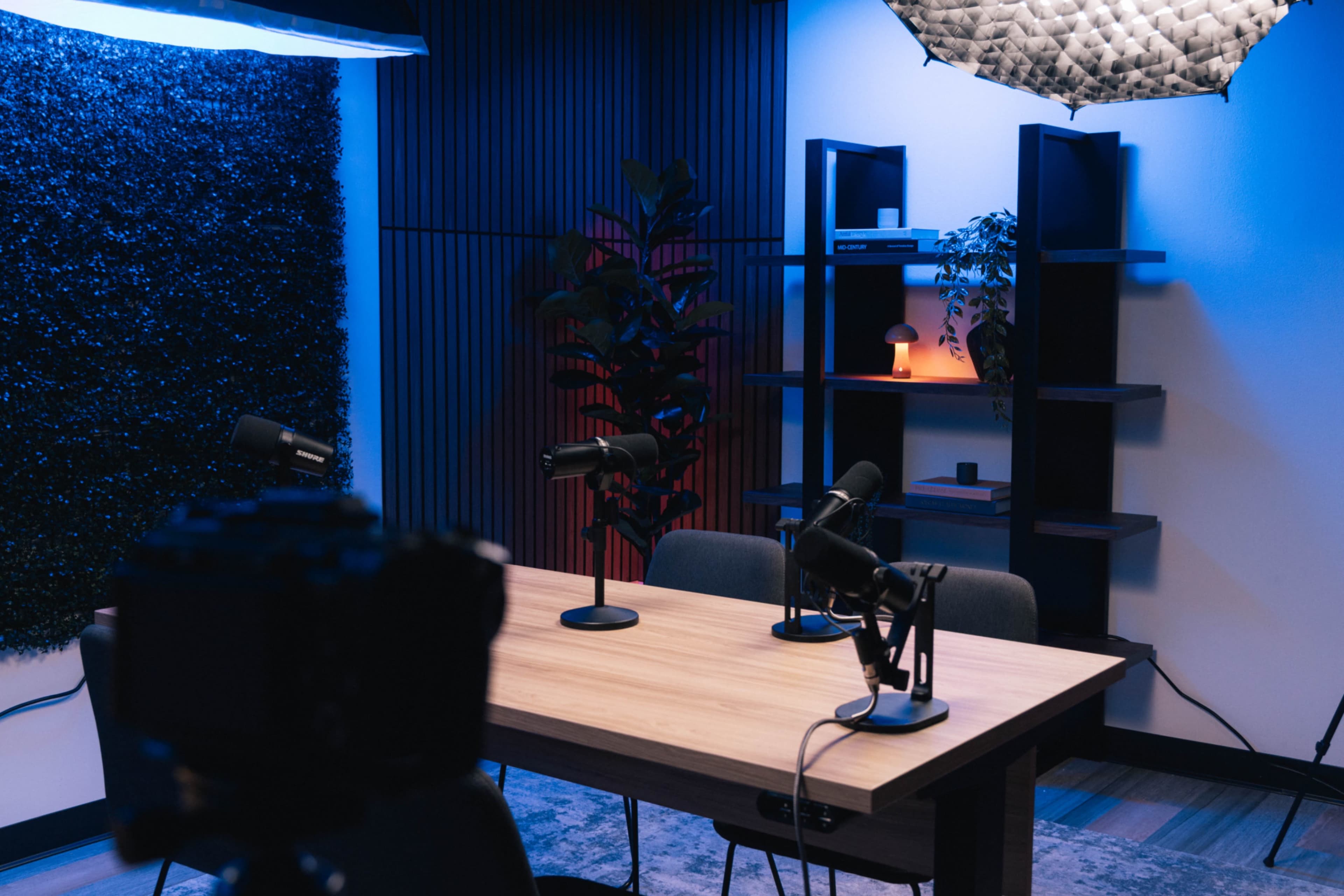The image shows a modern conference room setup with a wooden table, three microphones on stands, and a backdrop featuring a textured wall and shelves with decorative items under soft lighting.