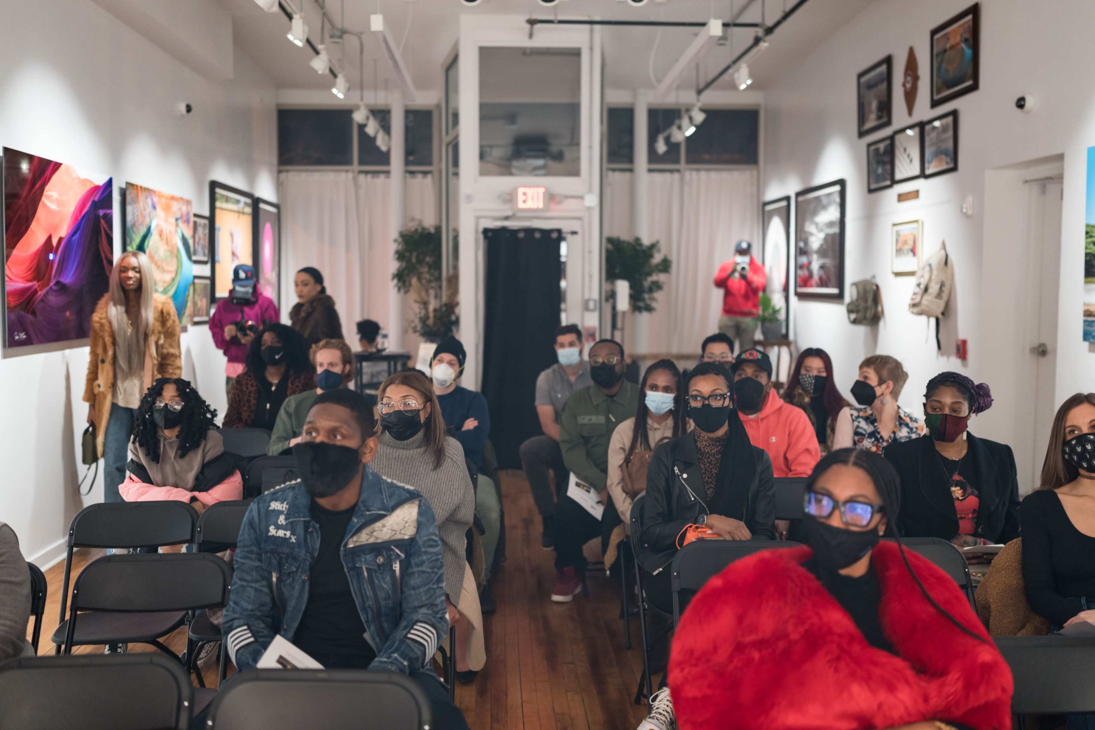 A group of people sit in an art gallery, wearing masks and looking towards the front of the room, which features various artworks on the walls.
