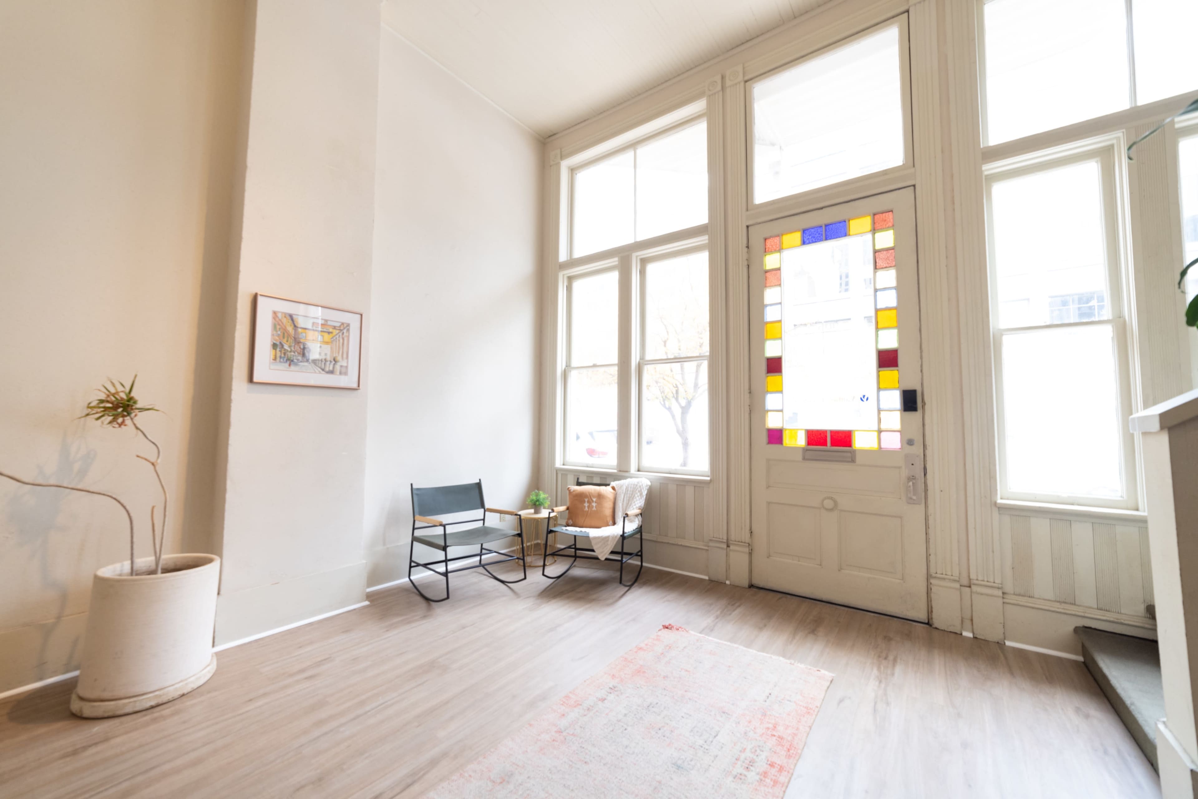 A bright foyer with large windows, a decorative door featuring colorful glass, a small seating area with a chair and a couch, a potted plant, and a light-colored wooden floor.