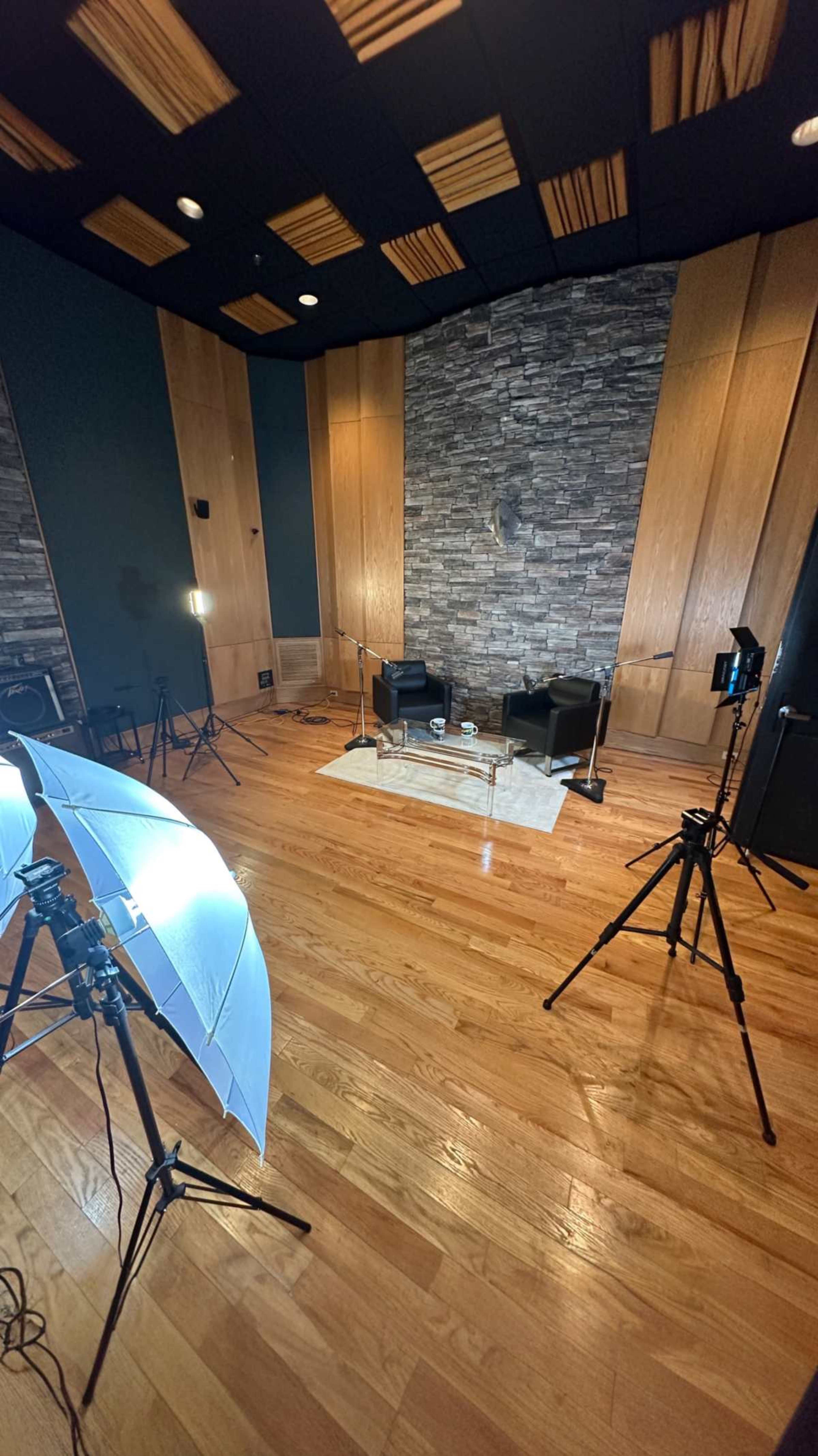 The image shows a well-lit studio space with two black chairs, a glass table, and multiple lighting equipment set up on wooden flooring against a backdrop of stone and wooden panels.