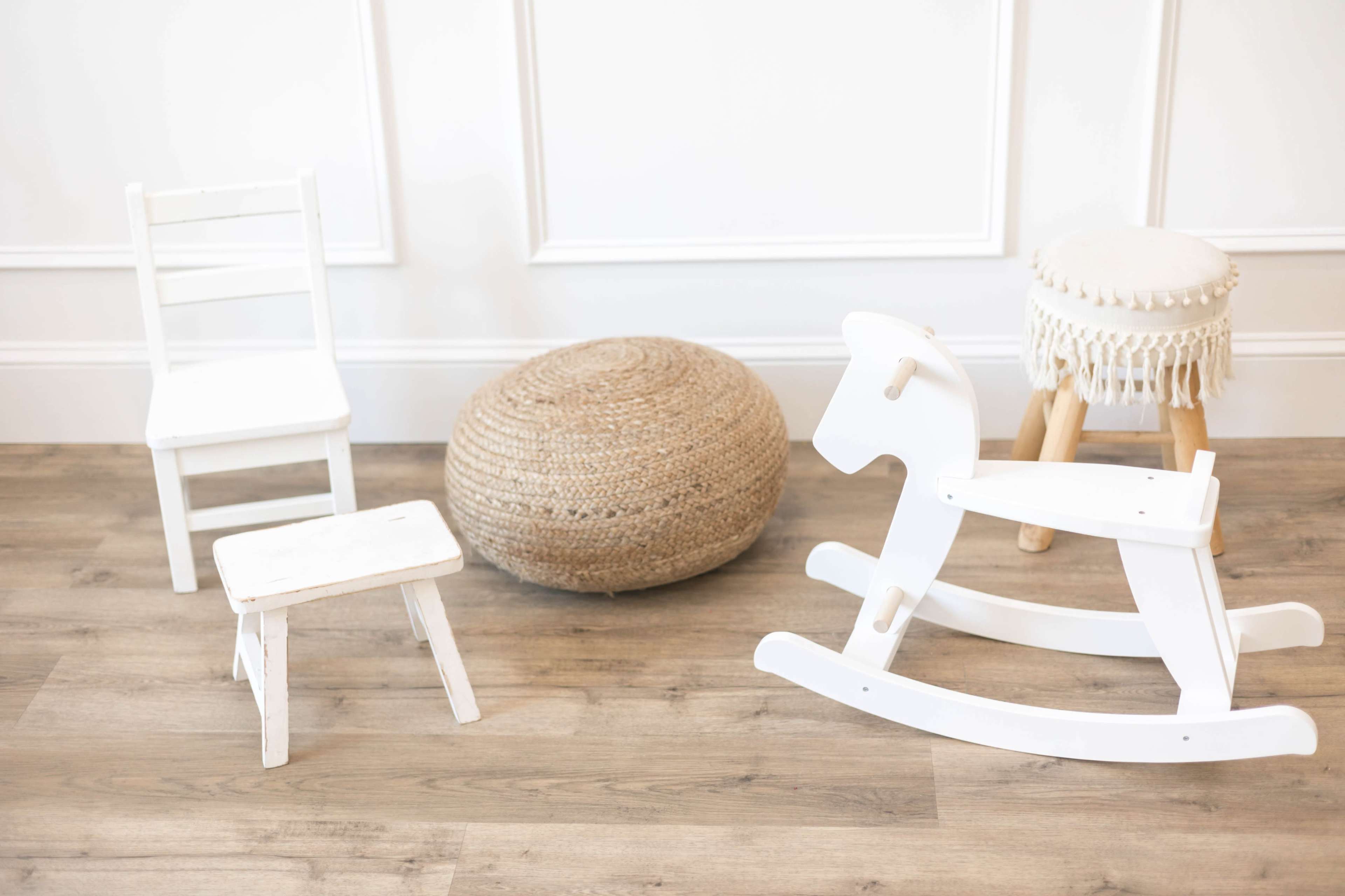 A white rocking horse, a small white stool, a white chair, and a woven pouf arranged on a wooden floor.