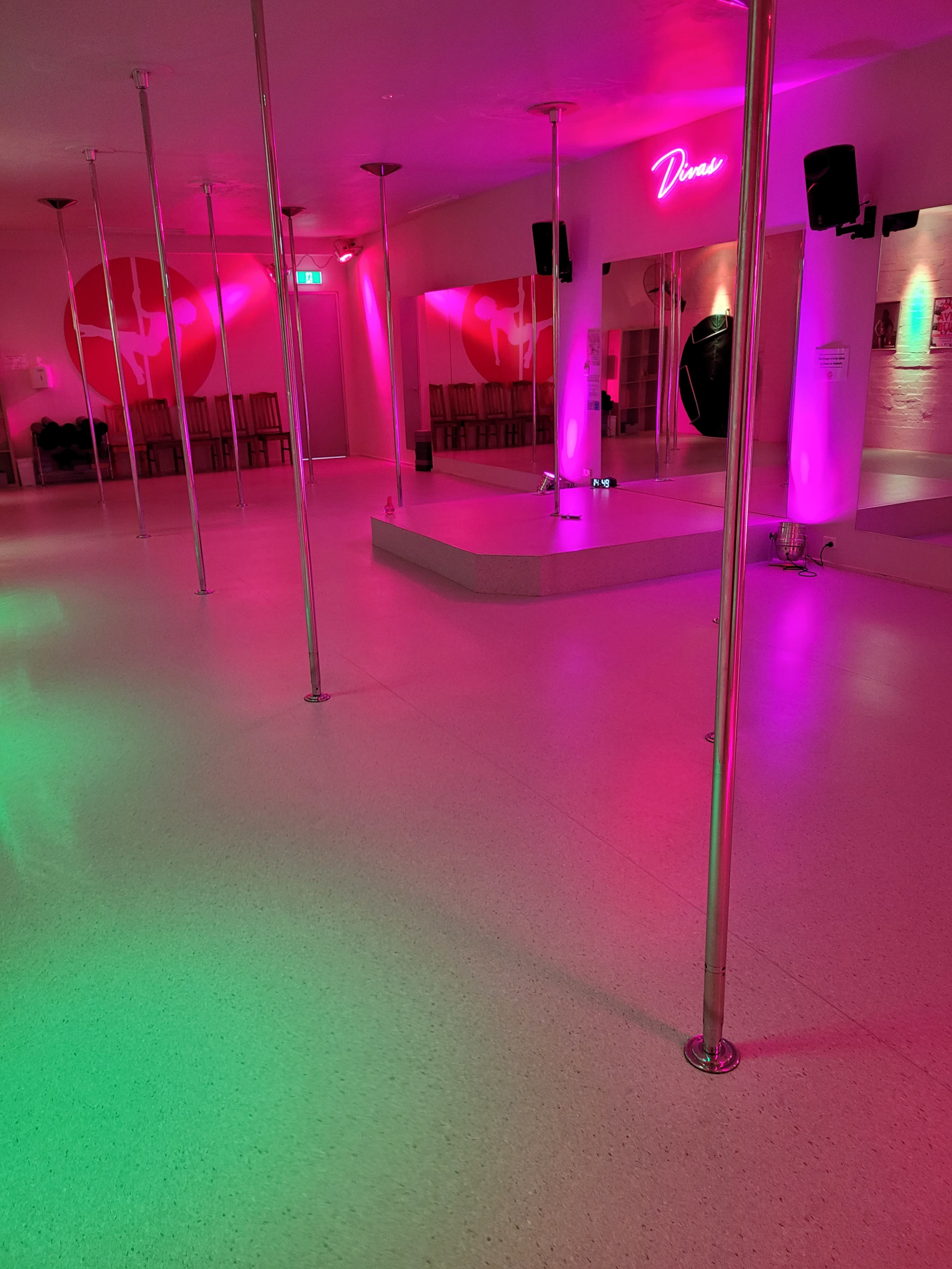 The image shows a dance studio with multiple vertical poles, illuminated by pink and green lighting, and a mirrored wall featuring the word "Dance."