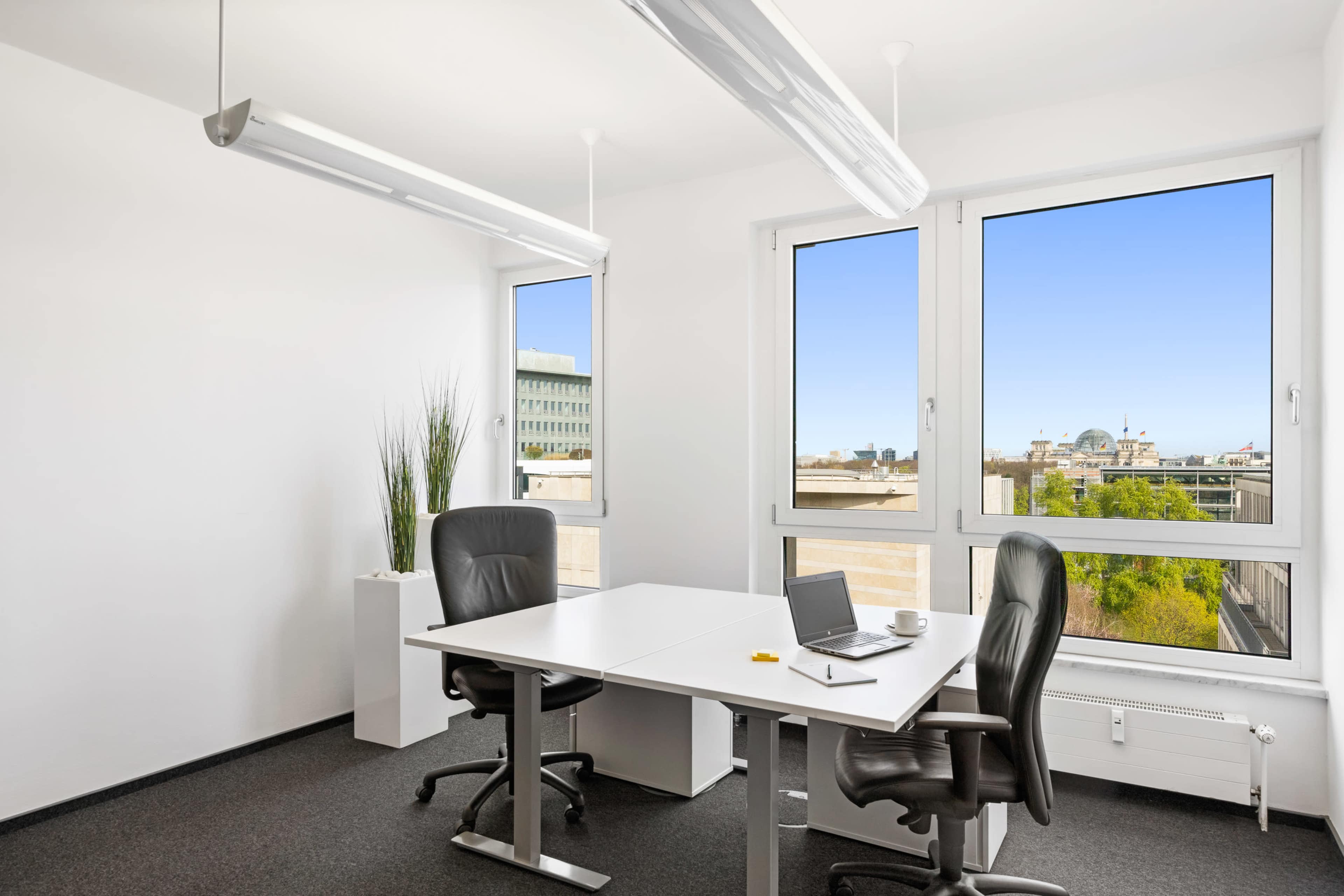 A modern office space features a white table with two chairs, a laptop, and large windows overlooking a cityscape.