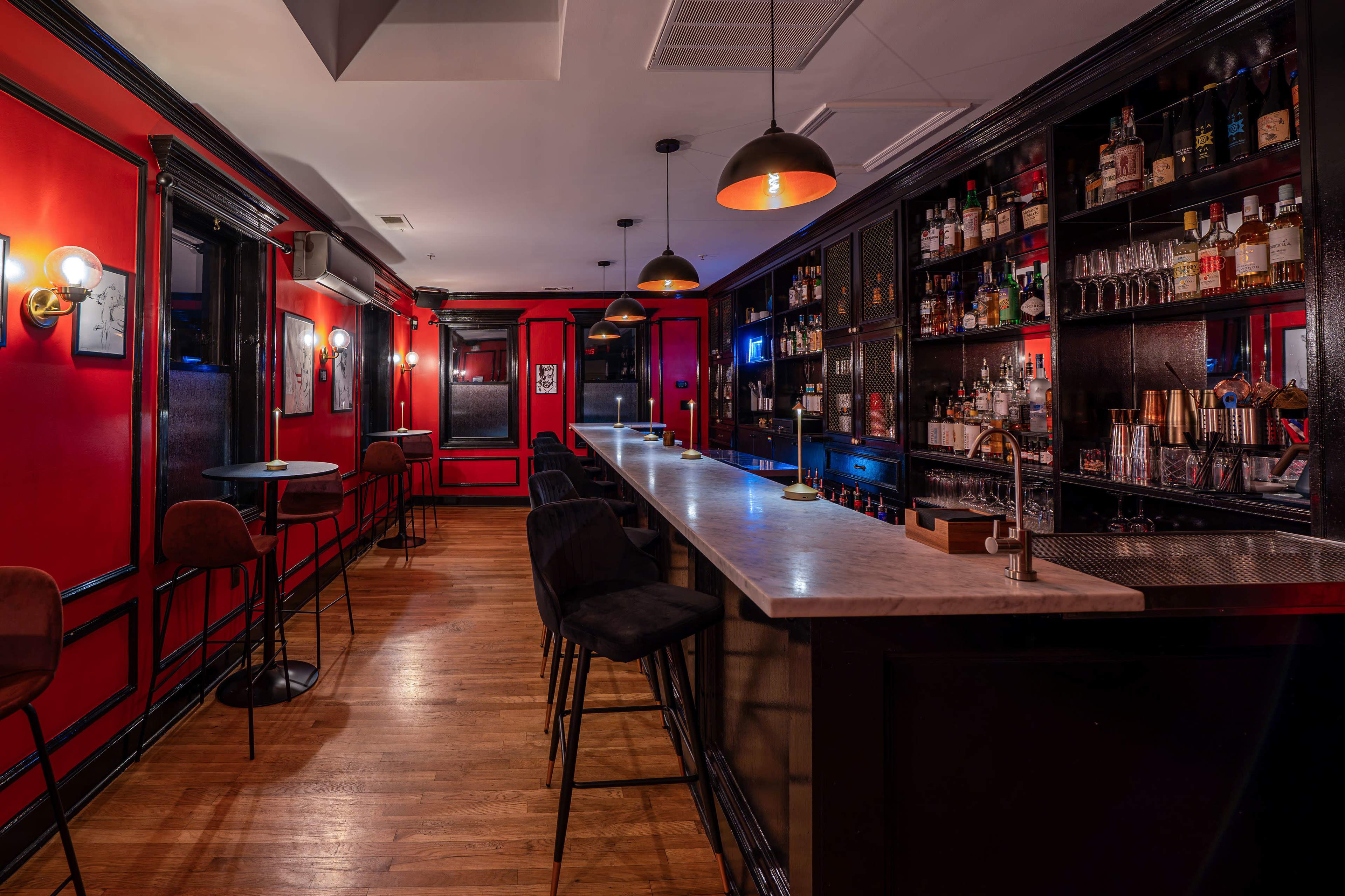 The image shows a dimly lit bar with red walls, a marble-topped counter, and shelves stocked with various bottles of liquor.
