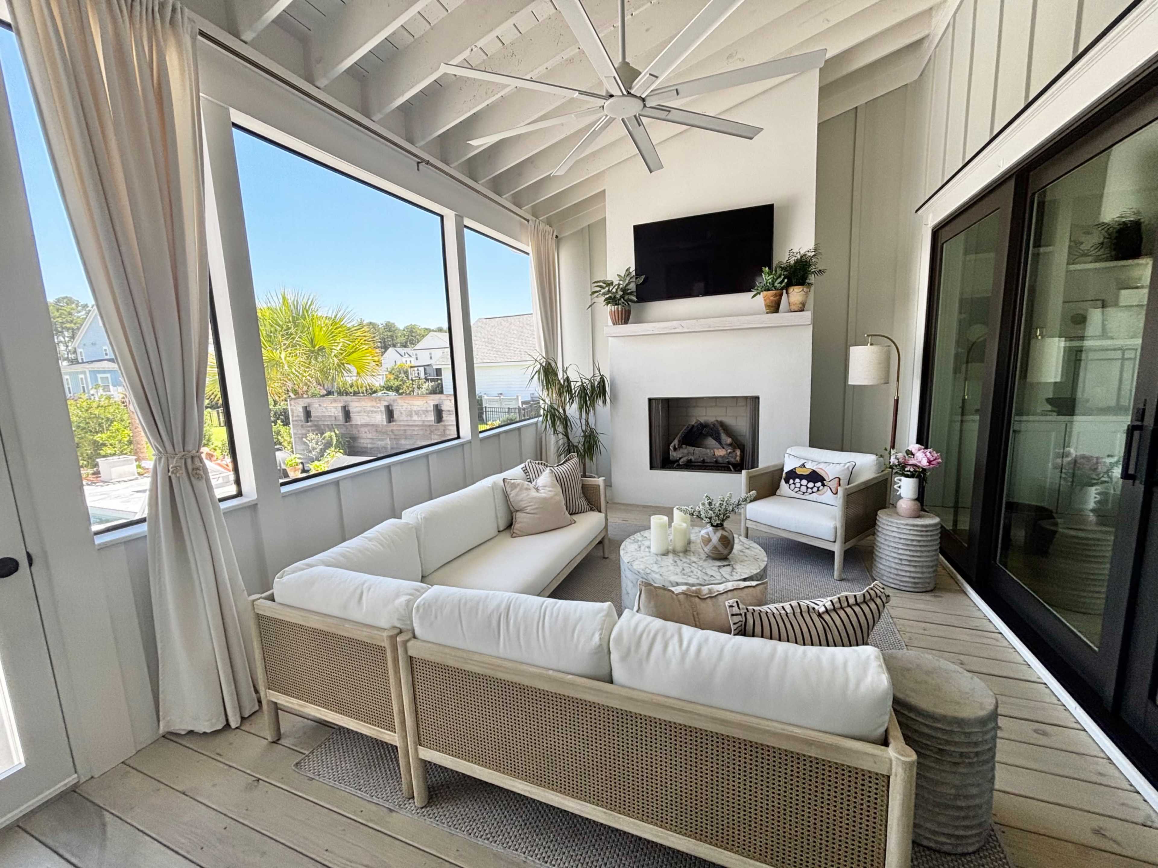 The image shows a cozy, modern sunroom with a white sectional sofa, a round coffee table, and large windows that provide a view of the outdoors.