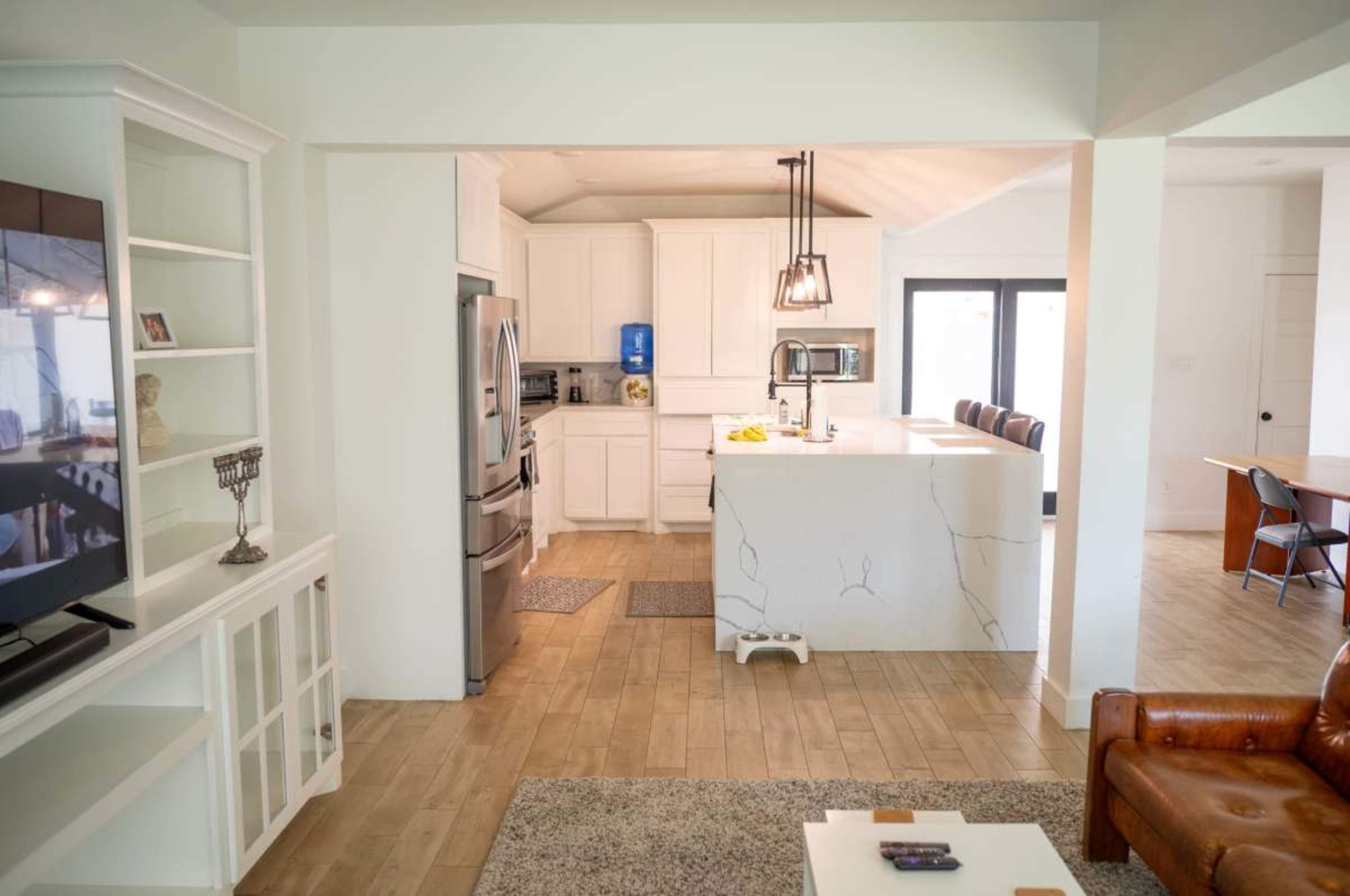 The image shows a modern open-concept living space with a kitchen, dining area, and living room, featuring white cabinetry, a large island, and hardwood floors.