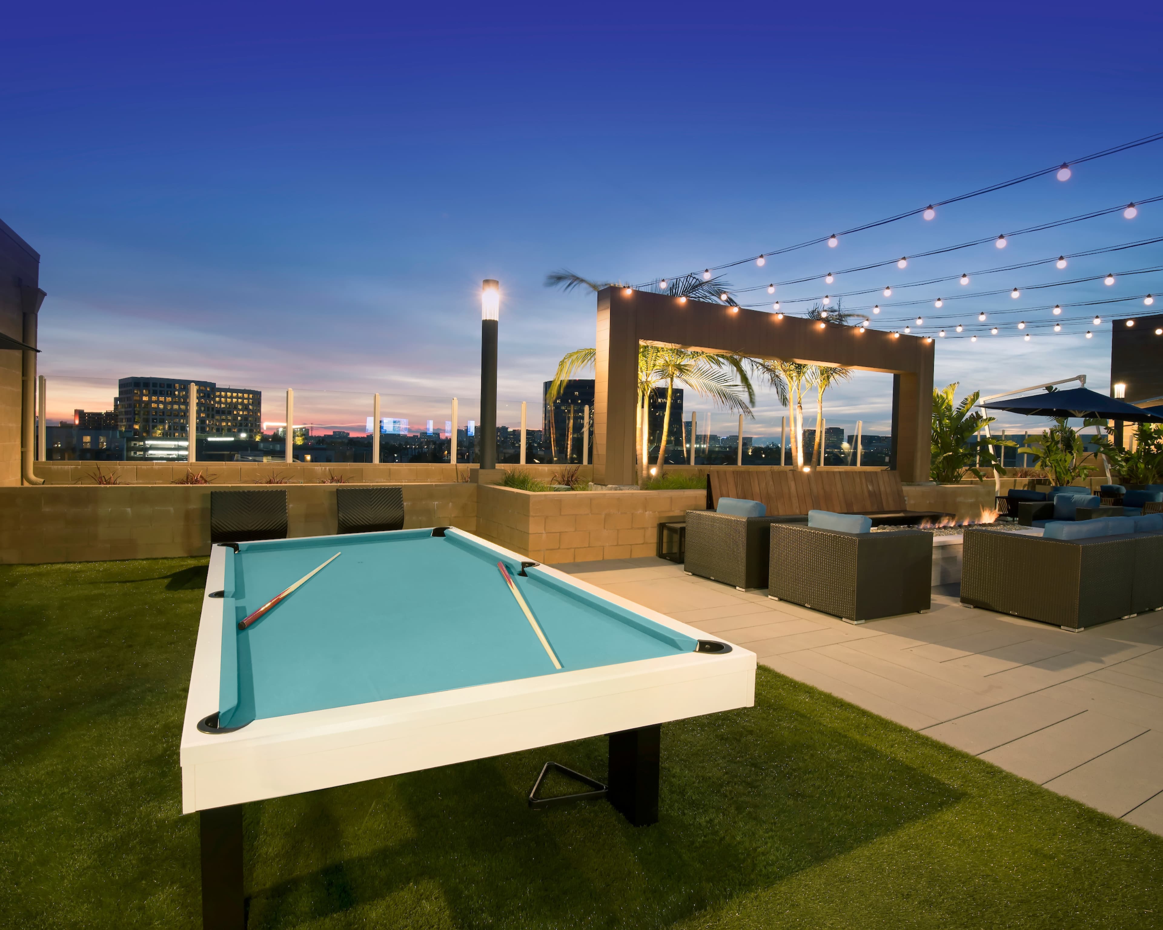 A rooftop terrace features a pool table surrounded by lounge seating and decorative string lights against a twilight sky.