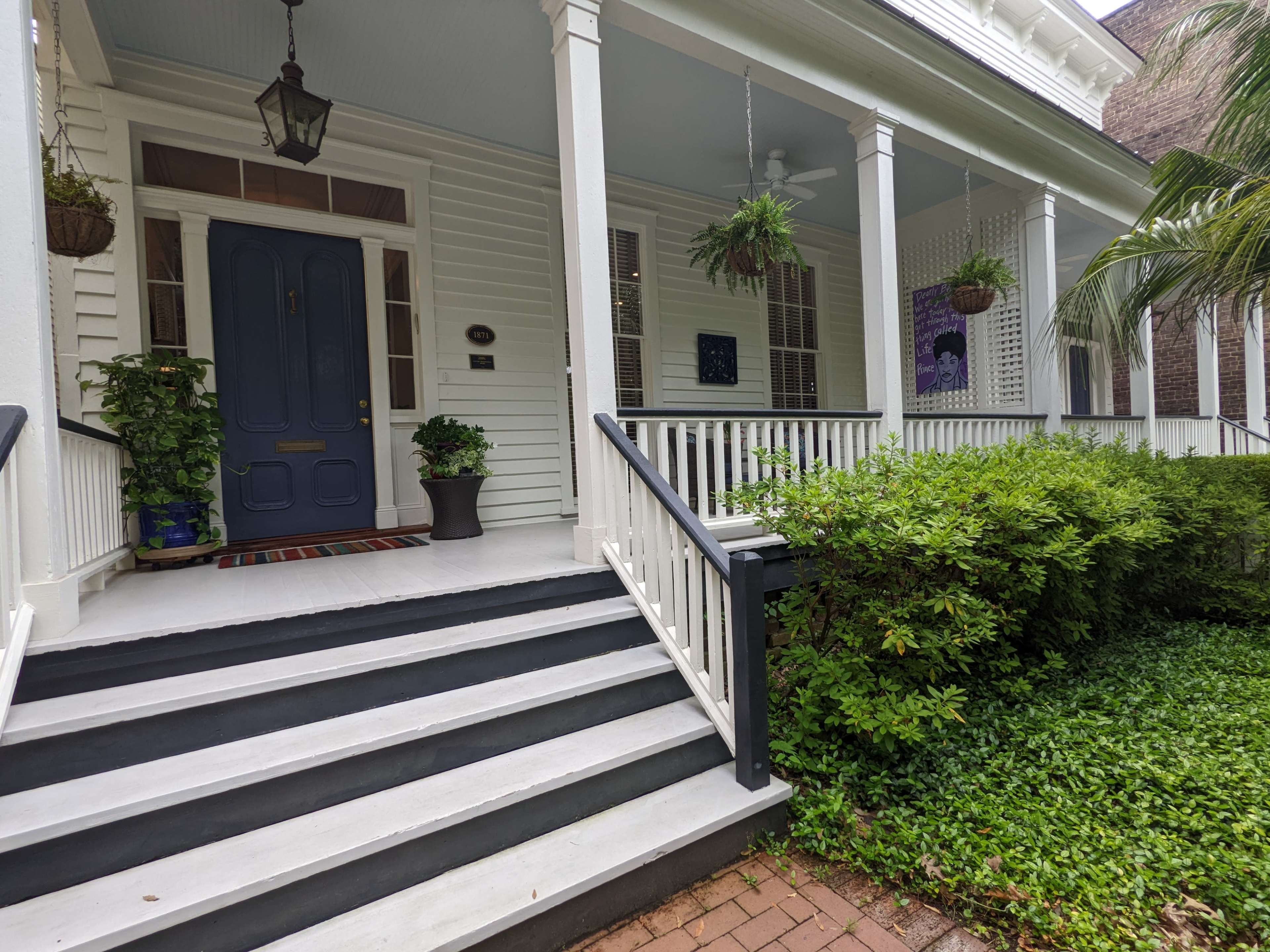 A white wooden porch with steps leading up to a blue front door, surrounded by green plants and a well-maintained garden.