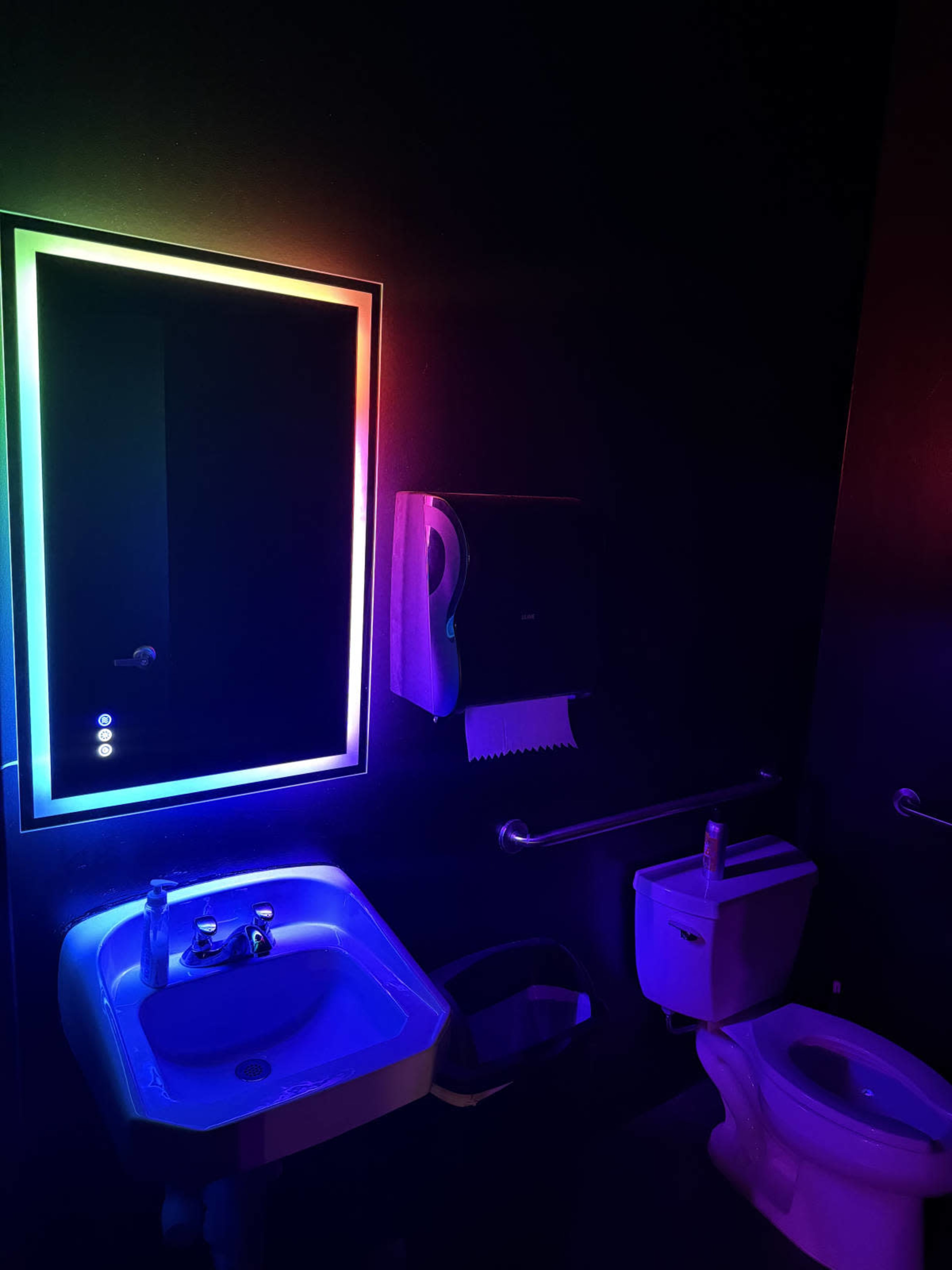 The image shows a bathroom with a glowing, multicolored mirror, a sink with a faucet, a toilet, and a paper towel dispenser against dark walls.