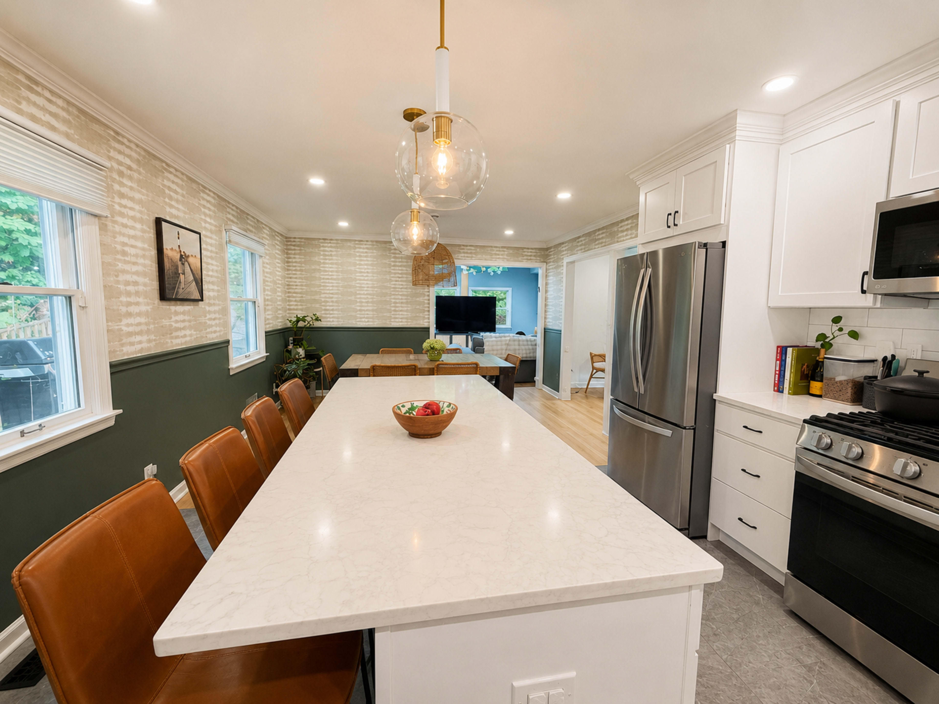 The image shows a modern kitchen featuring a long white countertop with brown leather bar stools, stainless steel appliances, and light green walls.