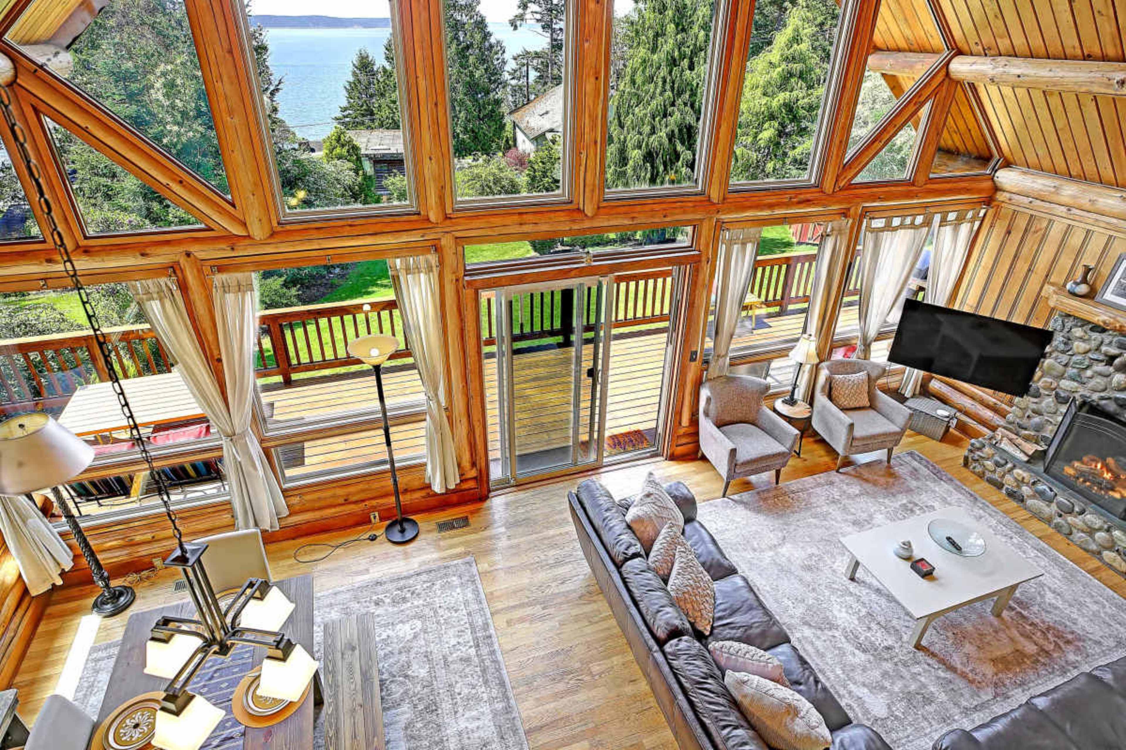 The interior of a spacious wooden cabin features large windows overlooking a scenic view, with a cozy living area furnished with a sofa and armchairs.