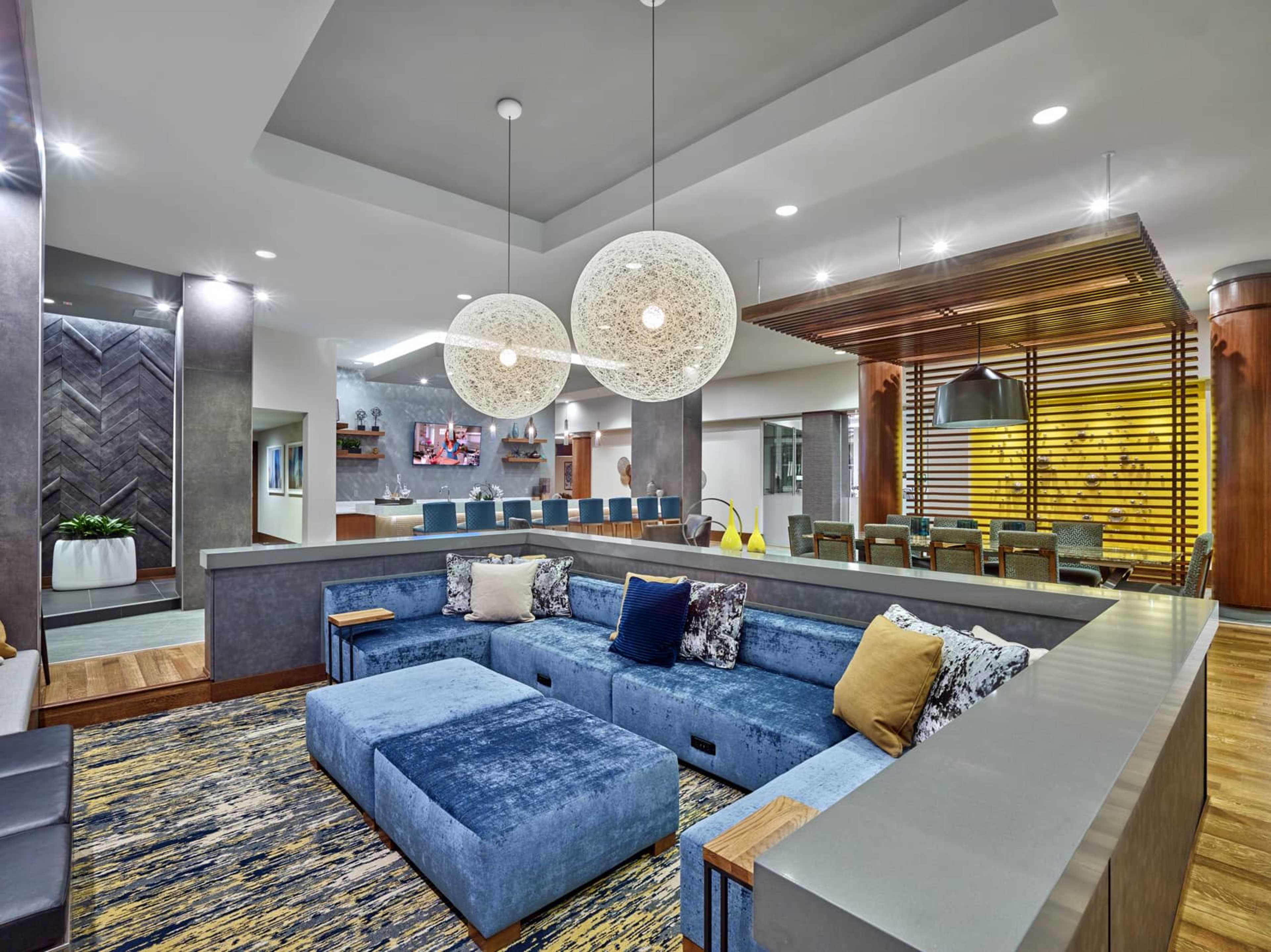 A modern lounge area featuring a blue sectional sofa, pendant lights, and a dining area in the background with wooden accents.