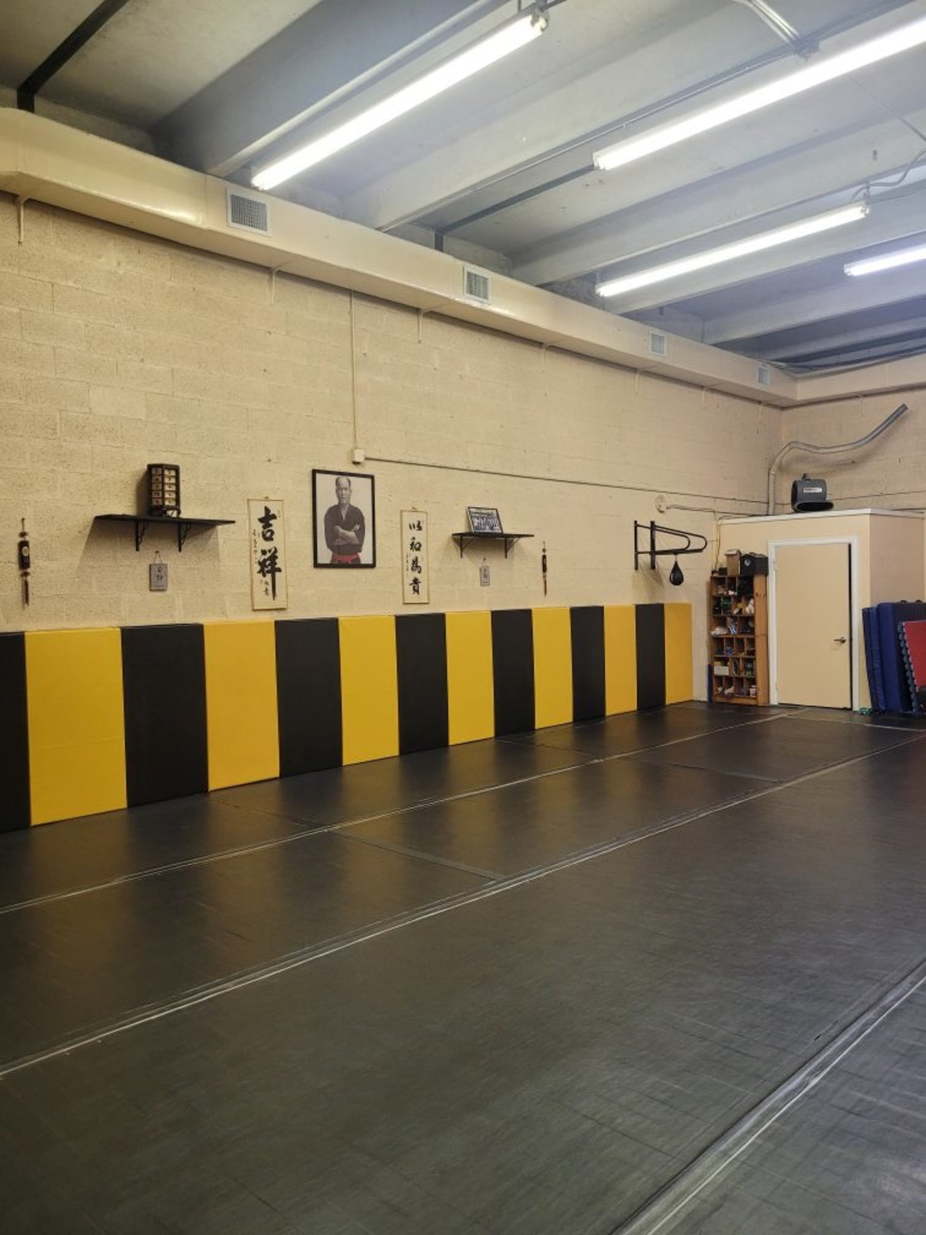 The image shows a martial arts training facility with black and yellow padded walls, a mat-covered floor, and a door leading to another area.