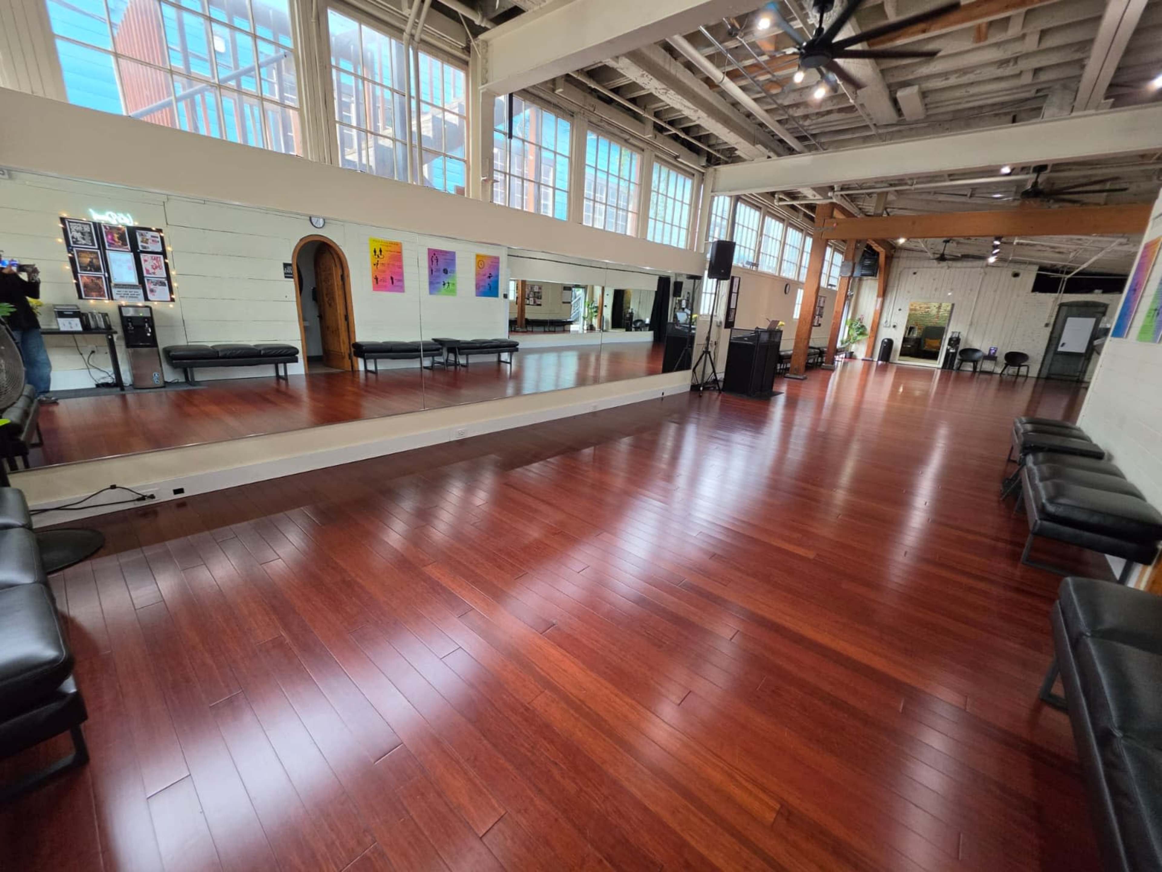 The image shows an empty dance studio featuring polished wooden floors, large mirrors, and ample natural light from the tall windows.