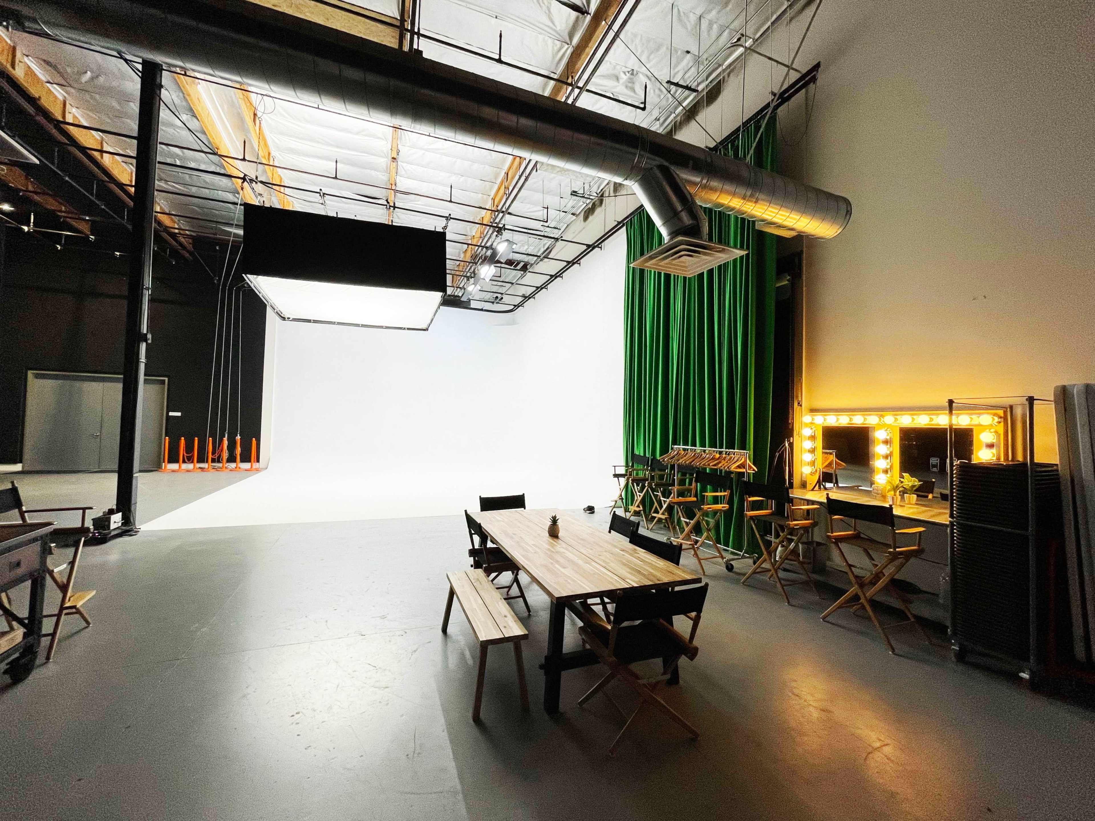 A spacious studio with a large white backdrop, green curtains, and various furniture including a wooden table and director's chairs.