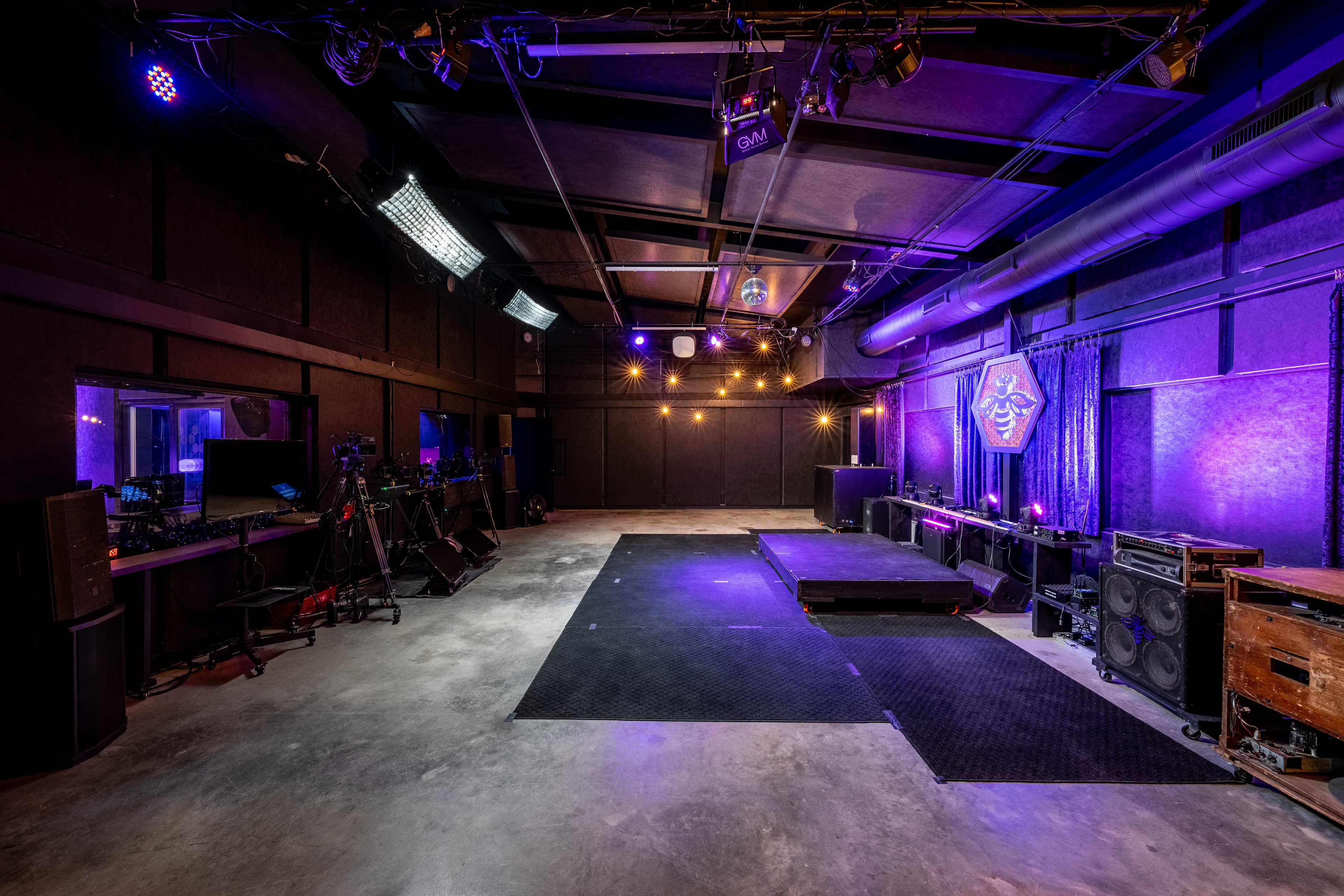 The image shows a spacious indoor venue with a performance stage, black walls, and dim lighting featuring purple and blue hues, along with sound equipment and monitors arranged along the sides.