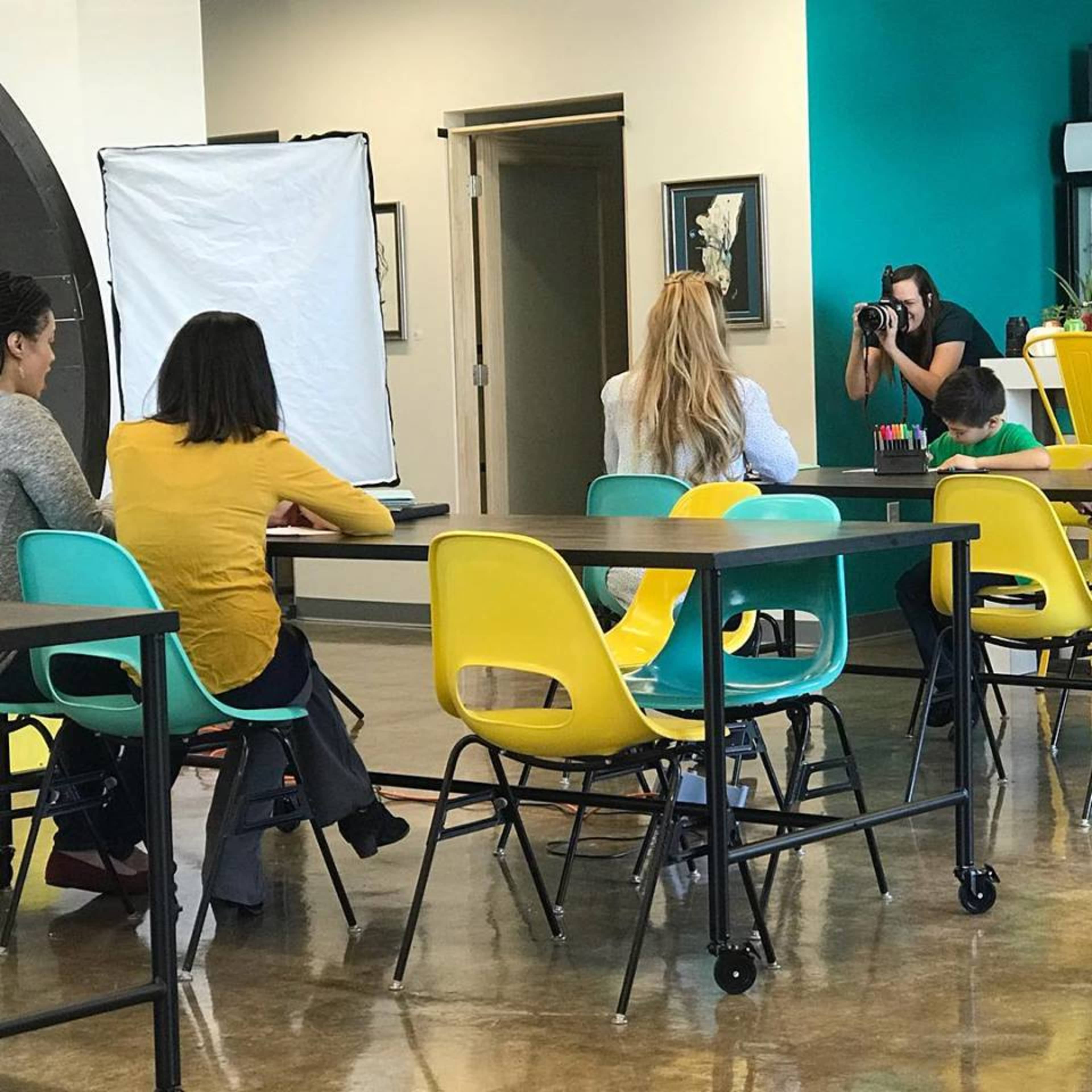 A group of individuals is seated at a table in an indoor space, with one person positioned in front of a camera being photographed.