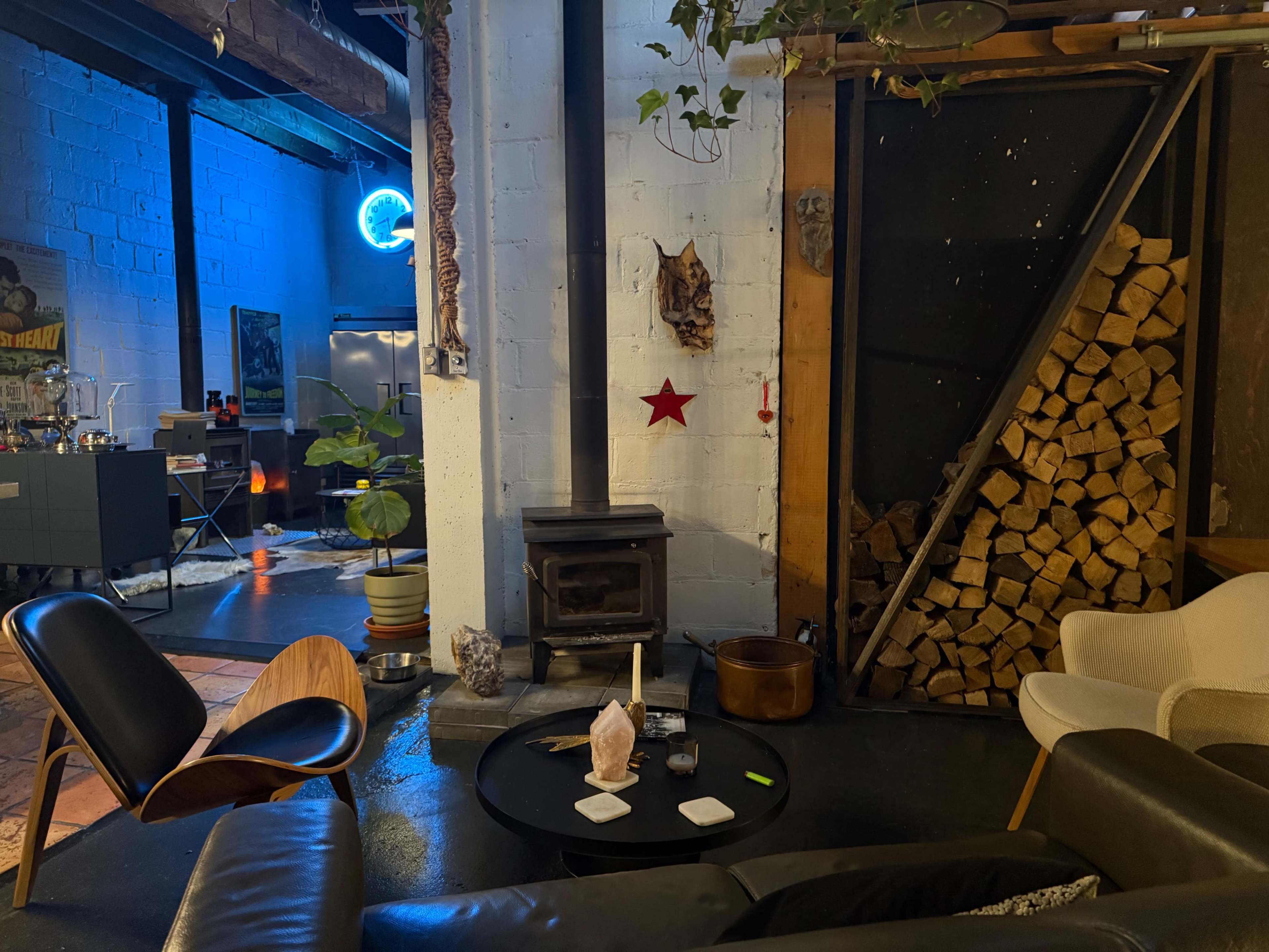 The image shows a cozy interior space featuring a wood stove, a stacked pile of firewood, a table with decorative items, and a mix of modern chairs.