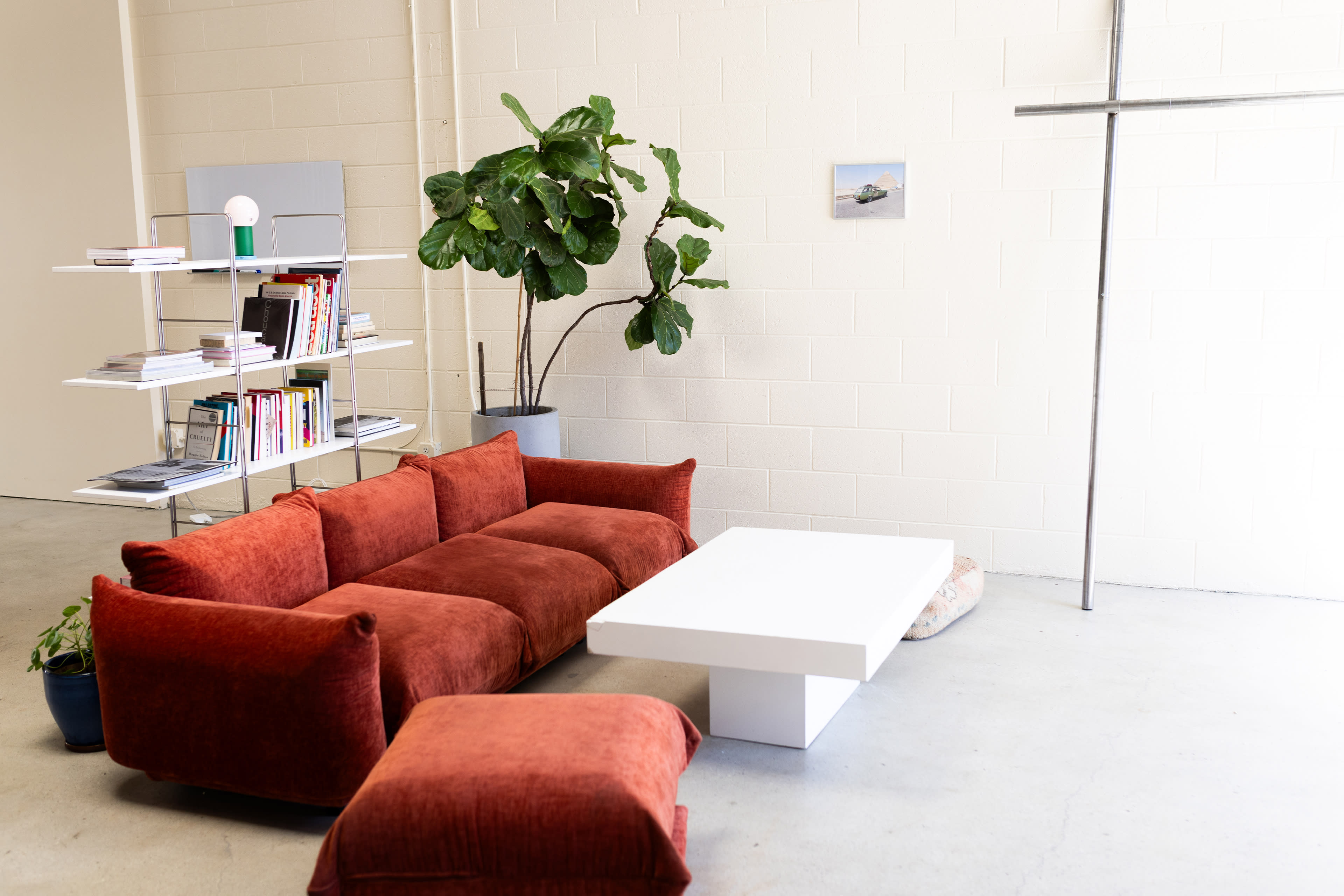 The image shows a modern interior space featuring a large orange sofa with a white coffee table, a potted plant, and a bookshelf against a light-colored wall.