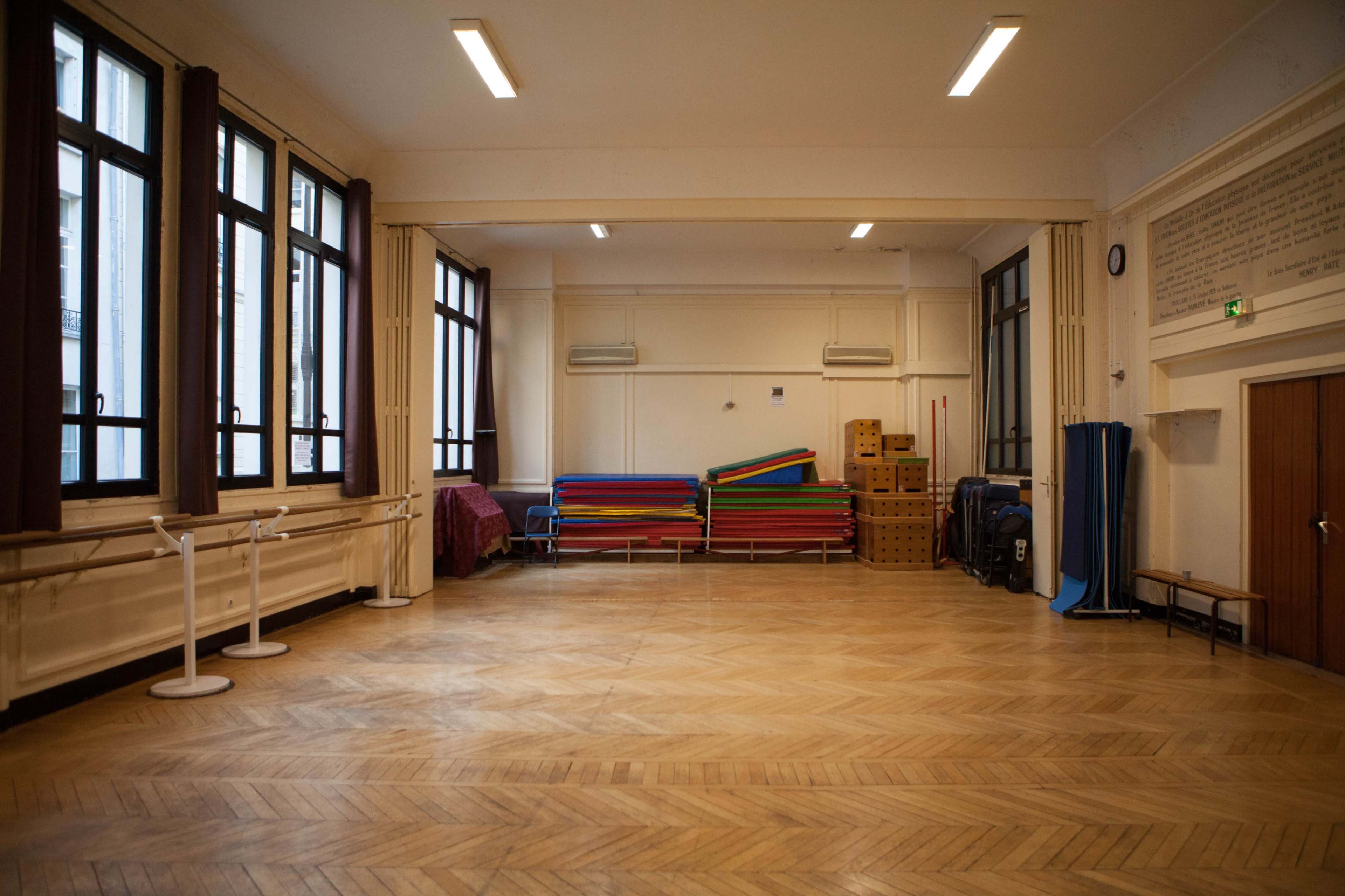 The image depicts a spacious, empty room with wooden flooring, large windows, and stacks of colorful mats in the corner.