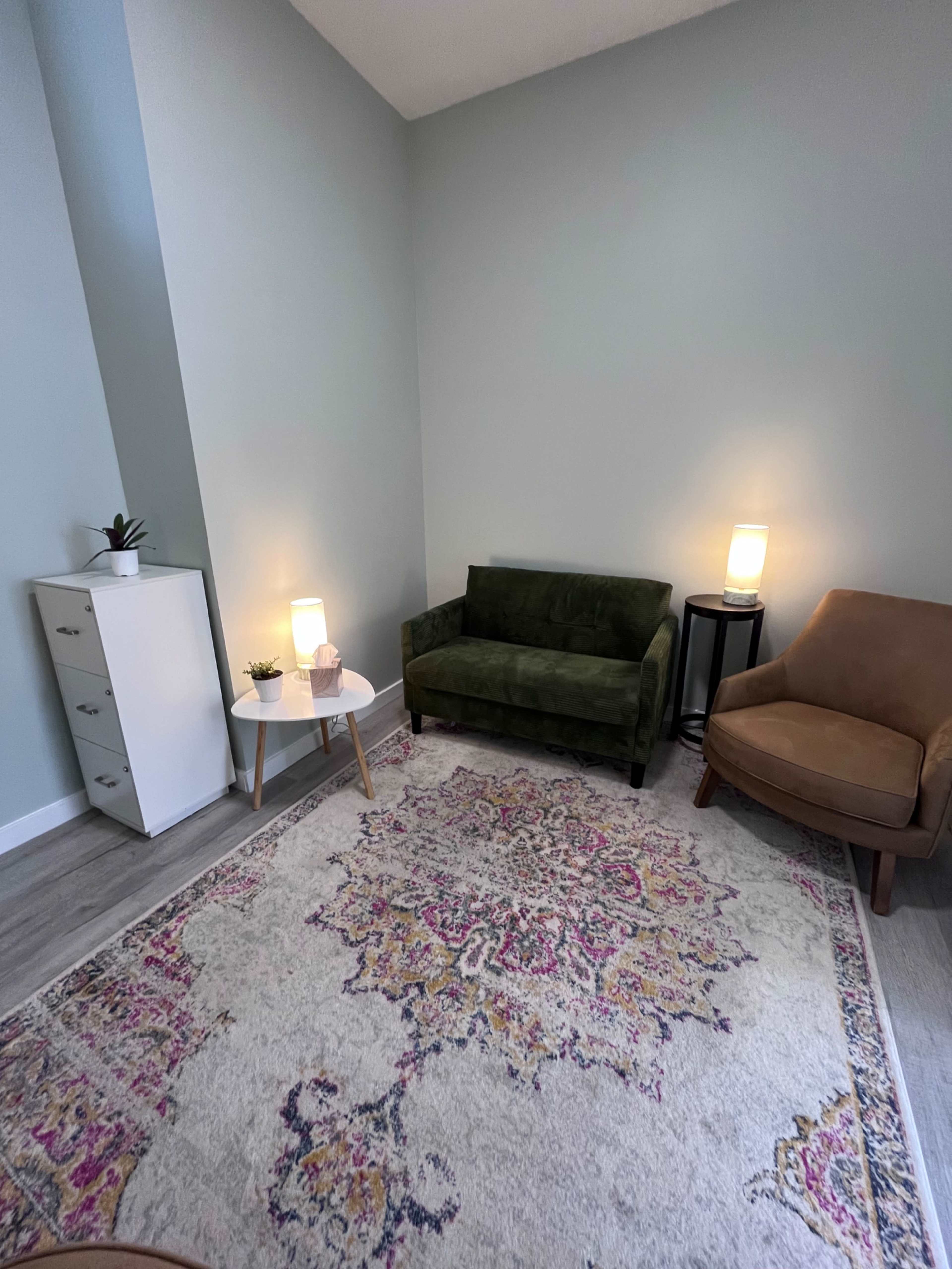 A small sitting area features a green sofa, a brown armchair, a round table with a plant, and two lamps on either side, all arranged on a decorative area rug.