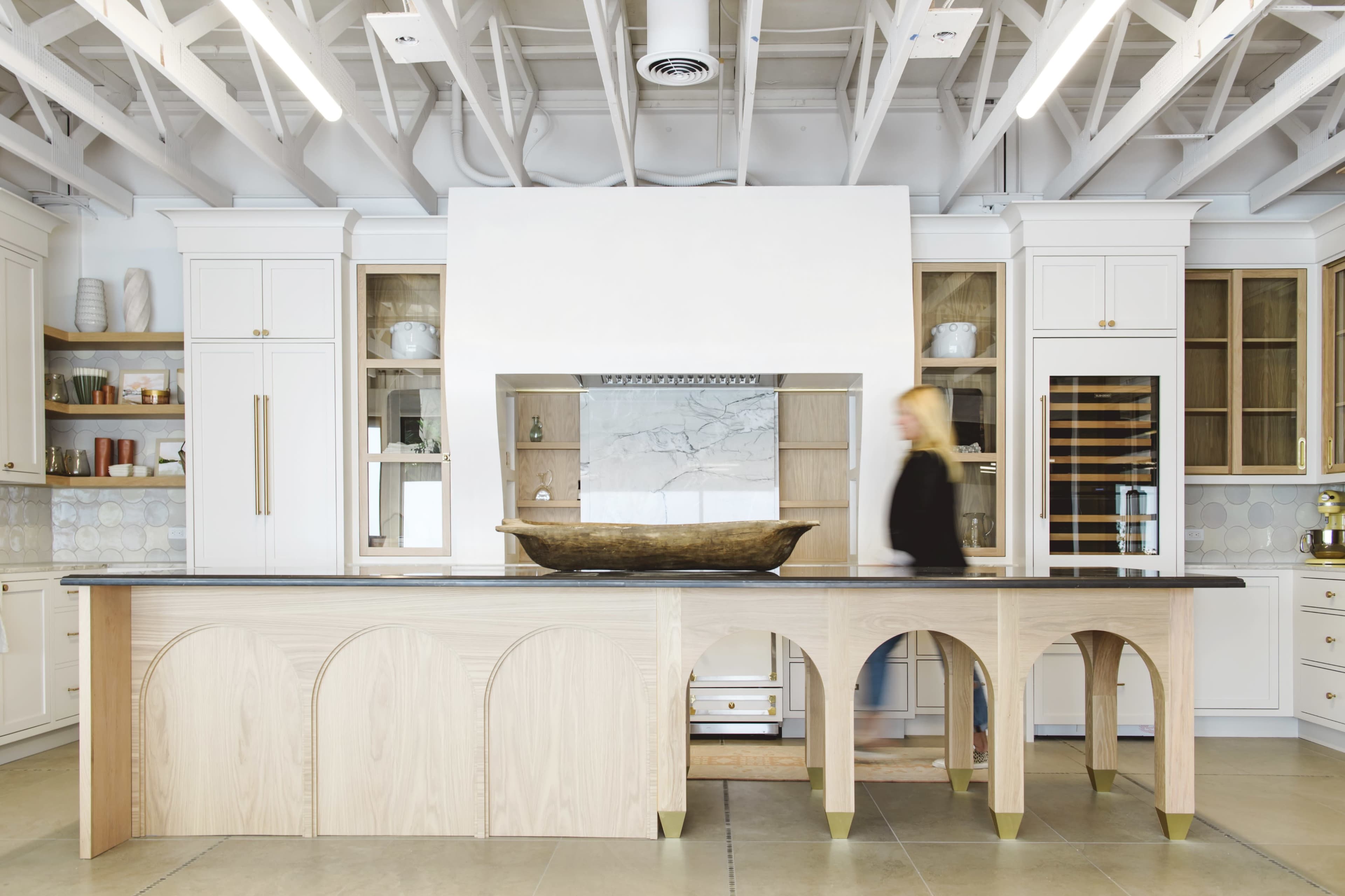 A modern kitchen features a large island with an elongated wooden bowl, white cabinetry, and a marble backsplash.