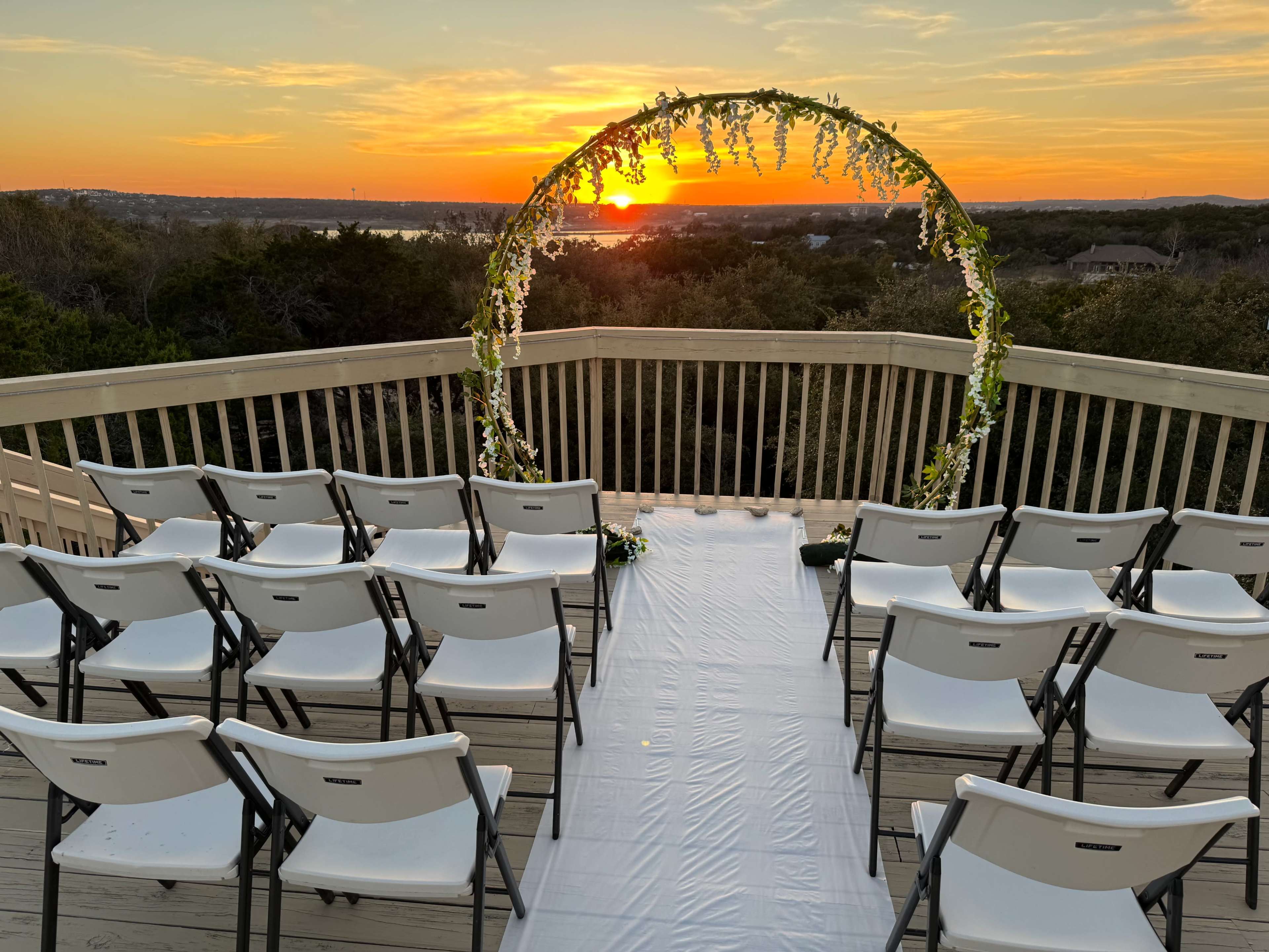 An outdoor wedding setup features white chairs arranged in rows facing a floral arch as the sun sets in the background.