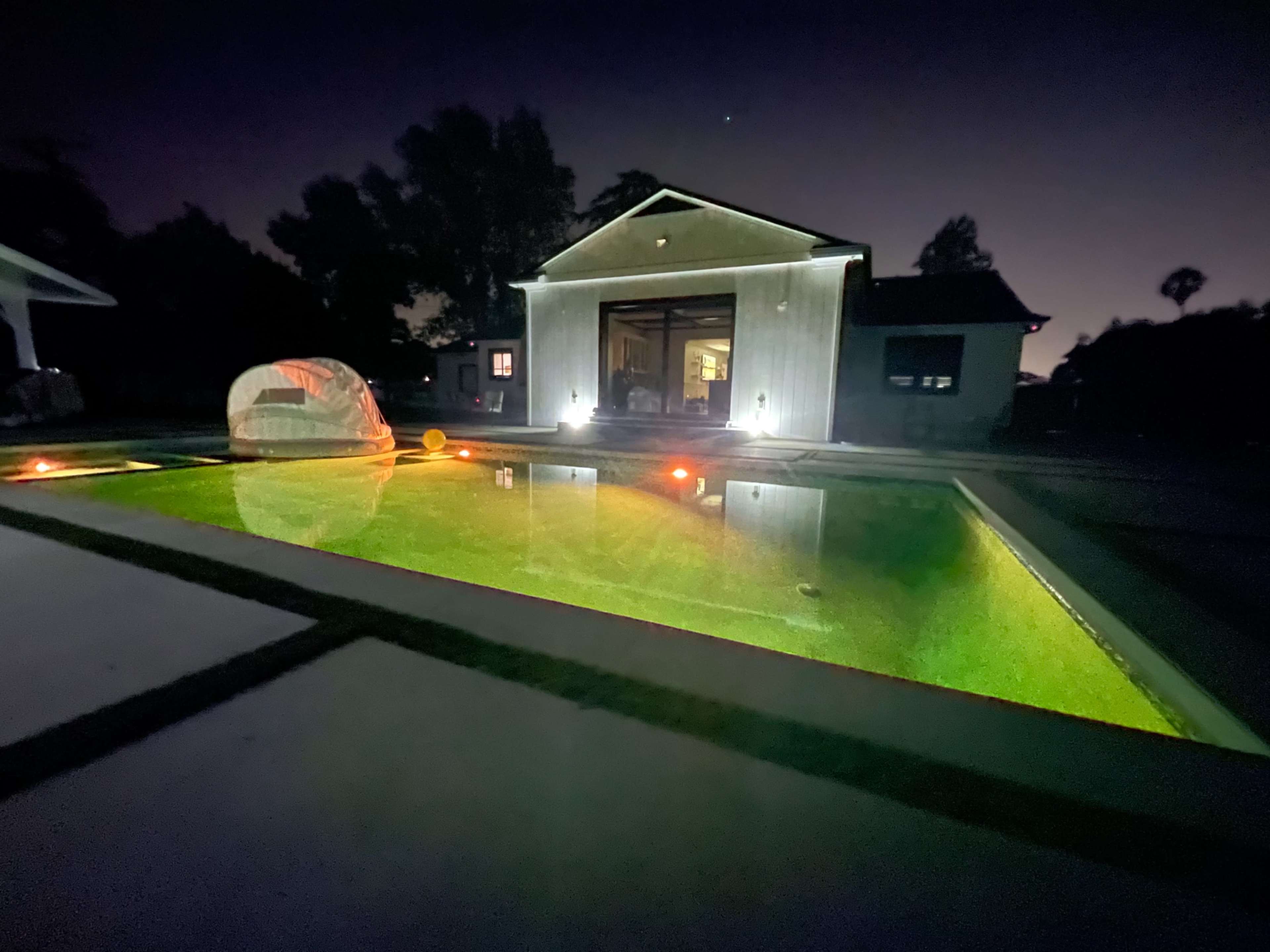 A dimly lit backyard pool area with a modern house in the background, reflecting green and orange lights on the water's surface.