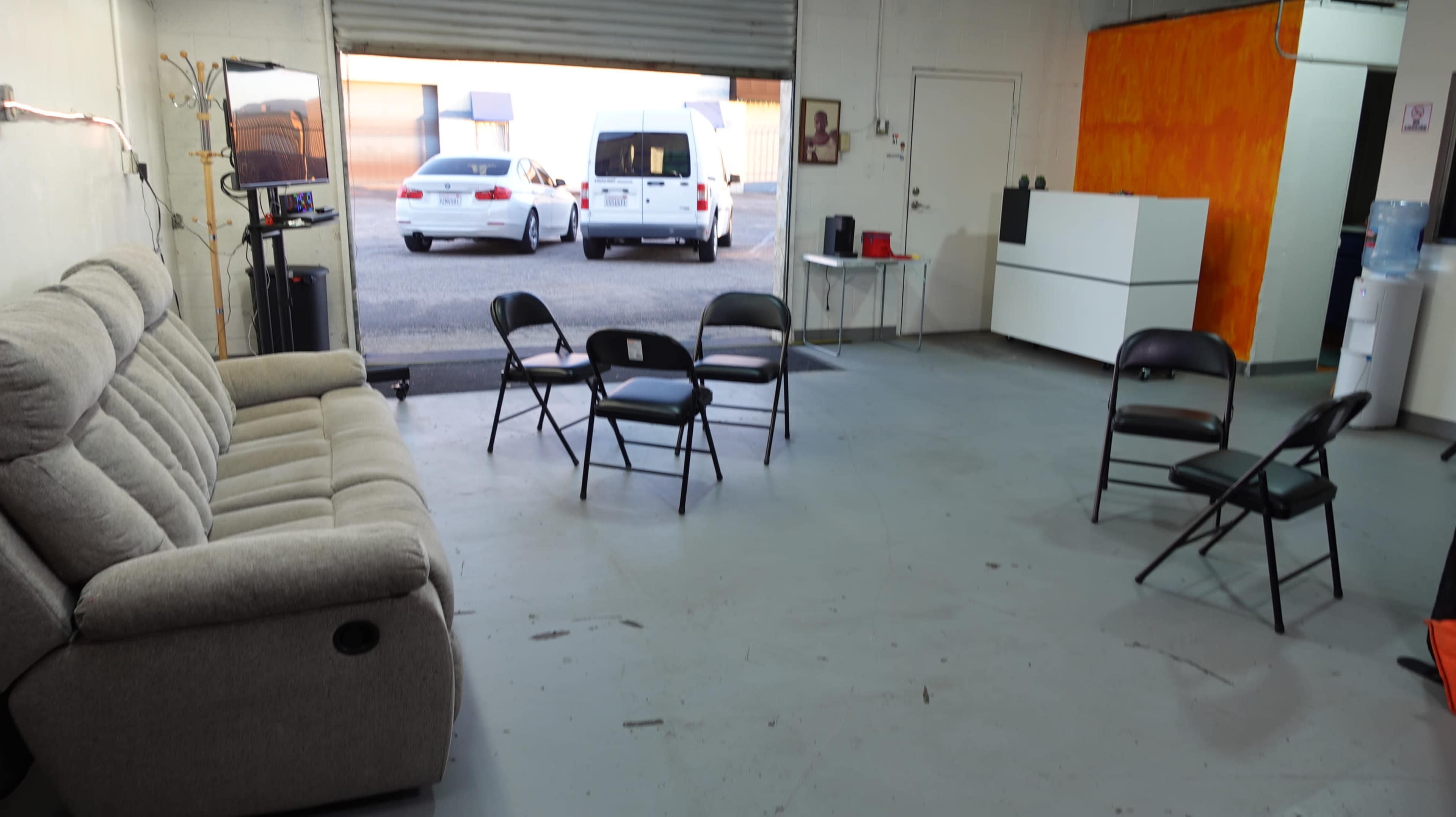 The image shows an interior space with a beige couch, several black folding chairs arranged in a circle, and a door leading to an outdoor area where two vehicles are parked.