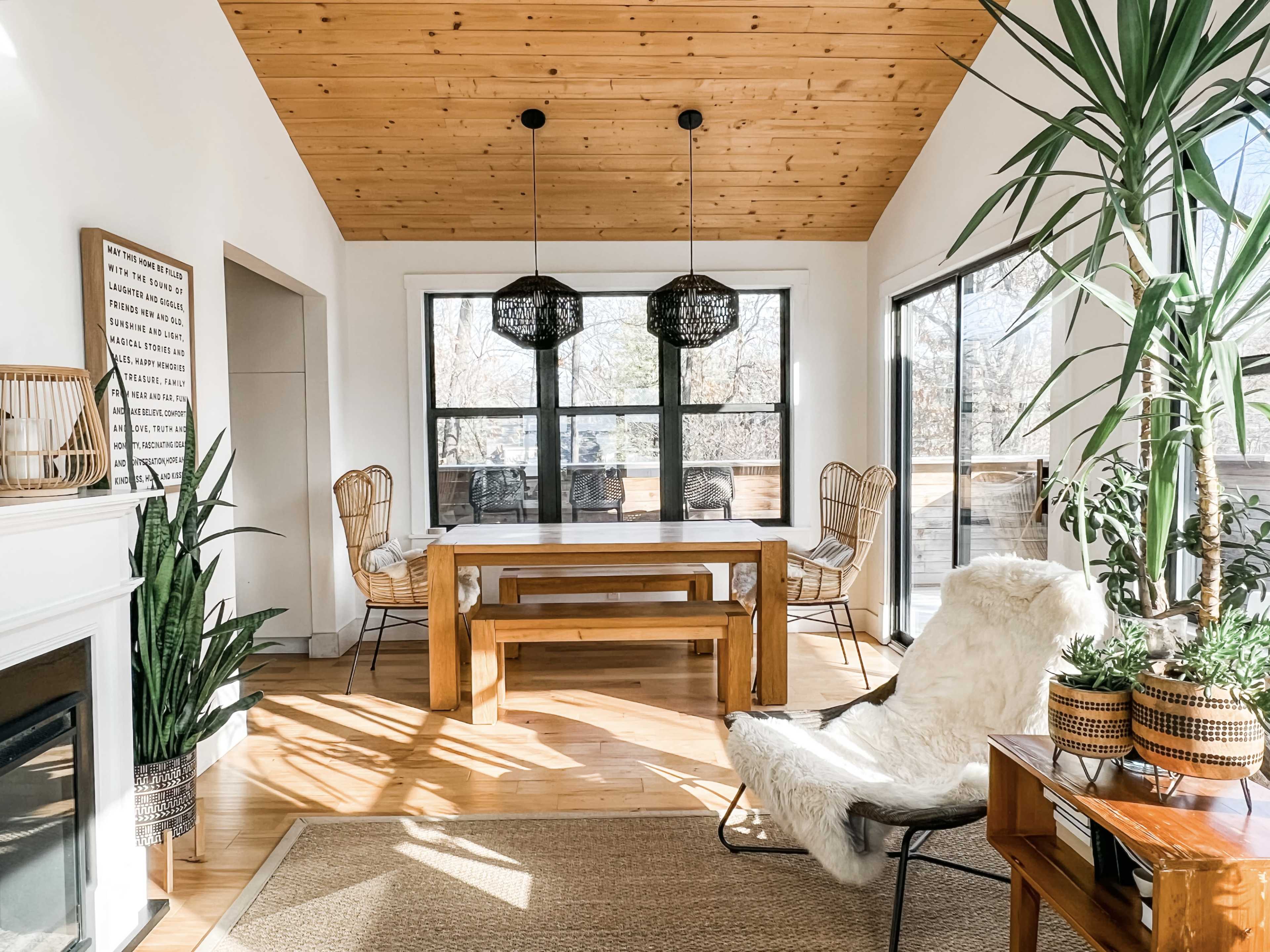 A dining area features a wooden table surrounded by chairs, large windows bringing in natural light, and a mix of indoor plants and decorative elements.