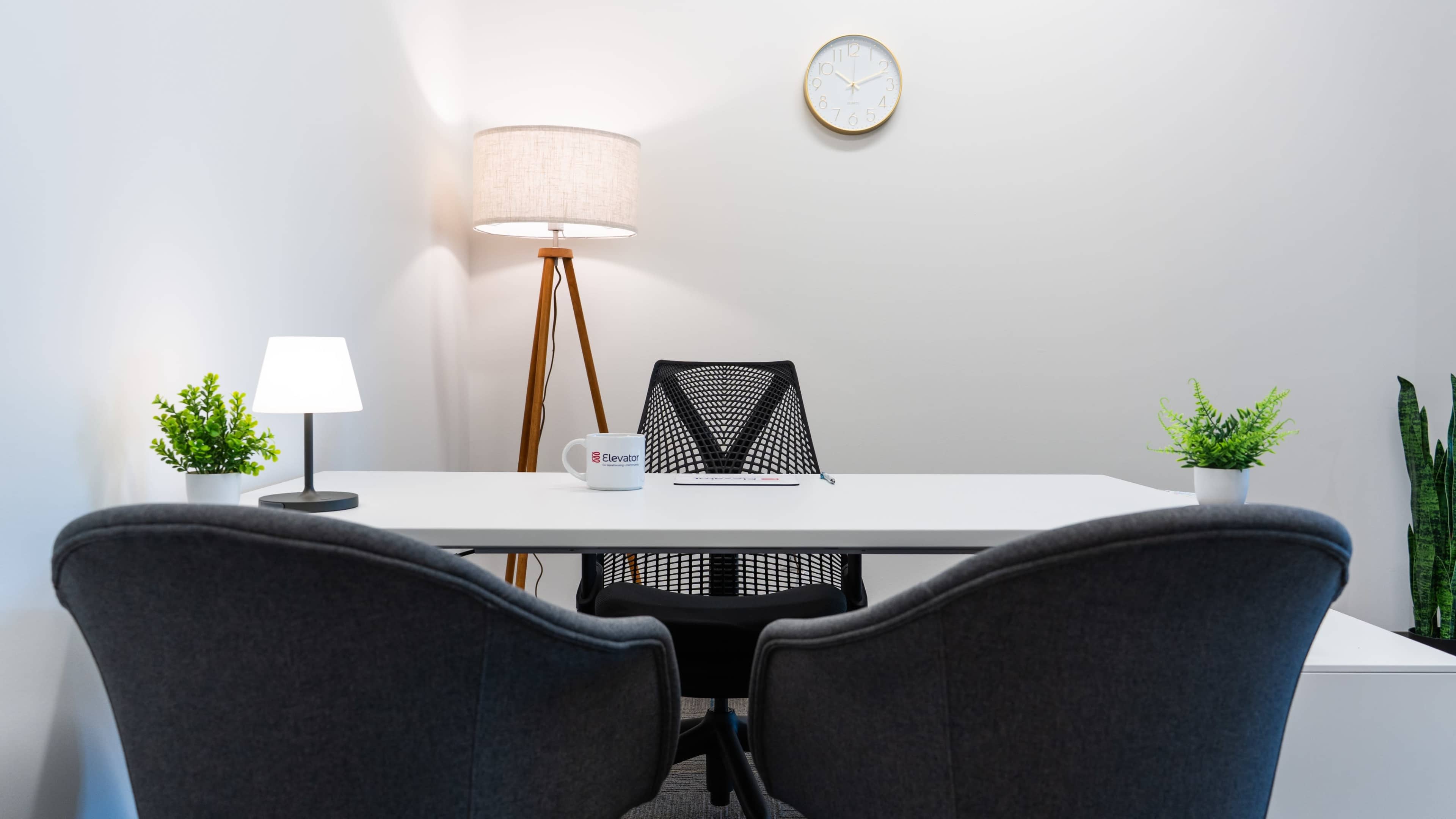 The image shows a modern office setup with a white desk, two gray chairs, a lamp, a clock on the wall, and small plants on the desk.