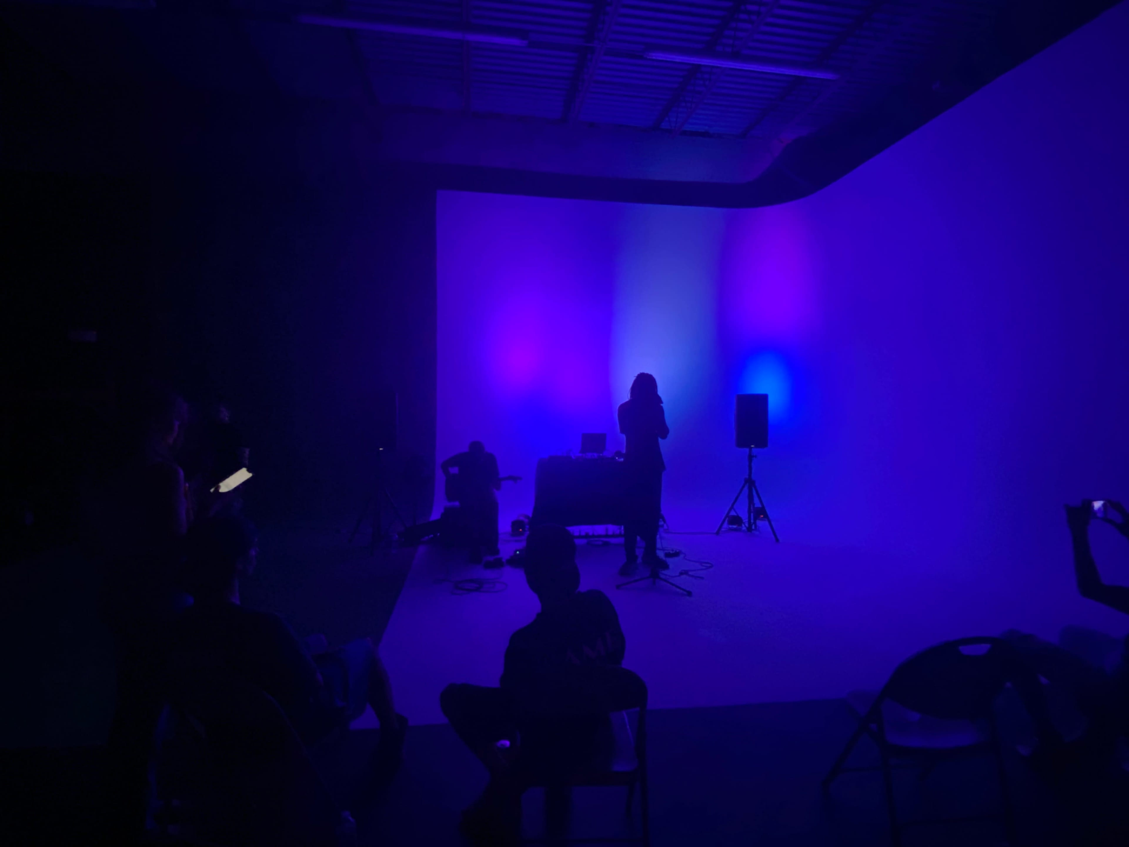 A person stands silhouetted against a backdrop of purple and blue lights while others sit in chairs observing the performance.